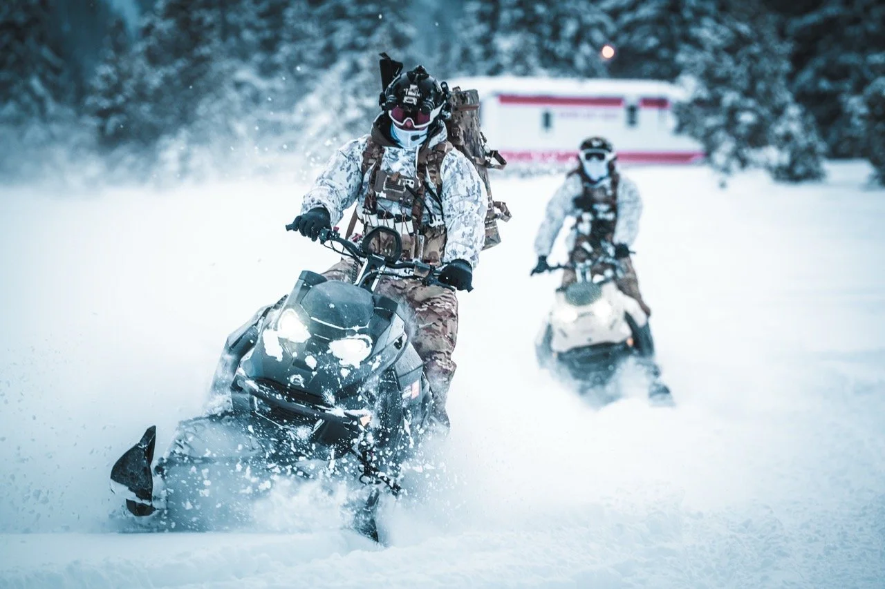 Two people riding snowmobiles through snow in a winter landscape with trees in the background.
