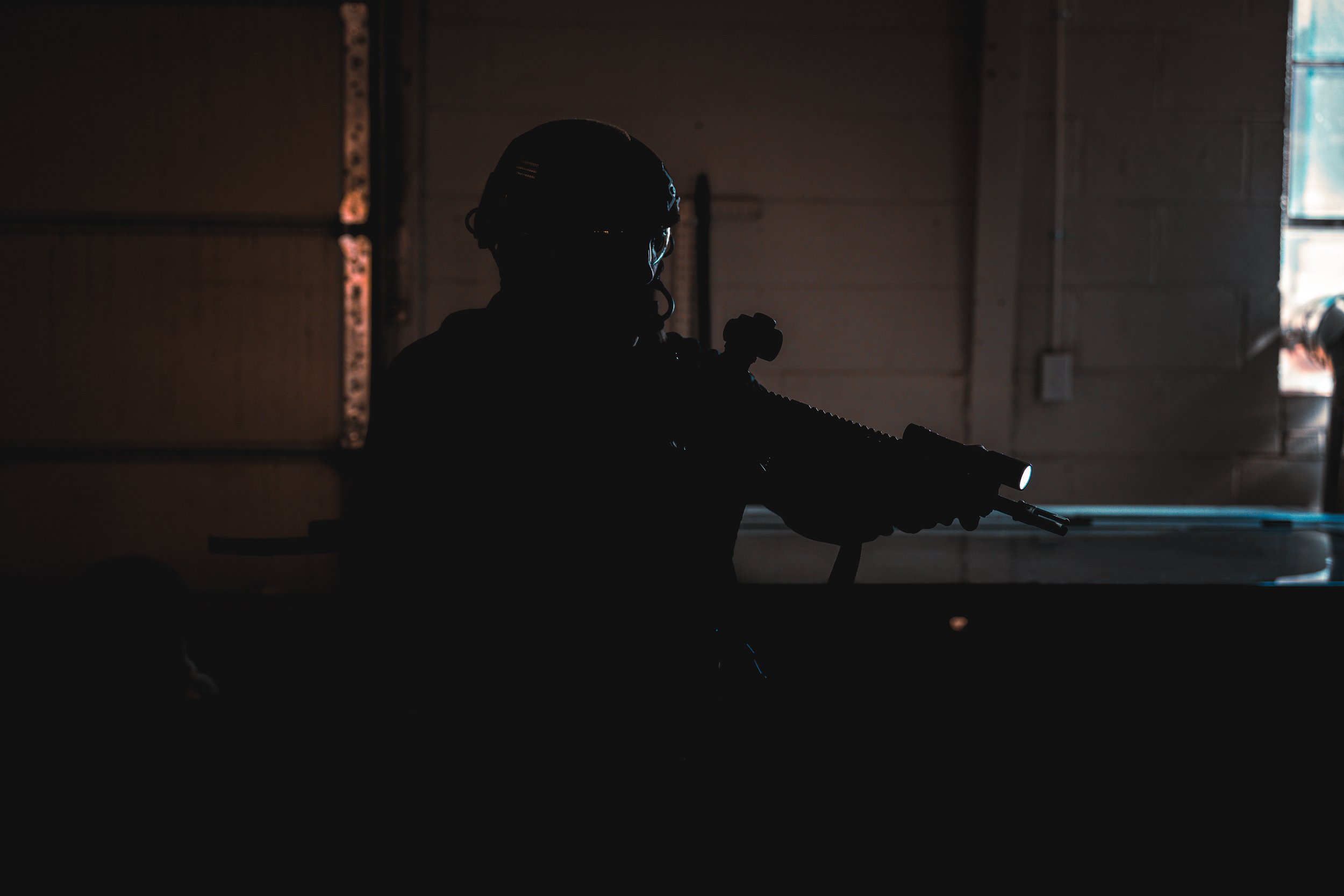 Silhouette of a person wearing a helmet and holding a rifle, with a faint light on the rifle's flashlight, inside a dimly lit garage or warehouse.