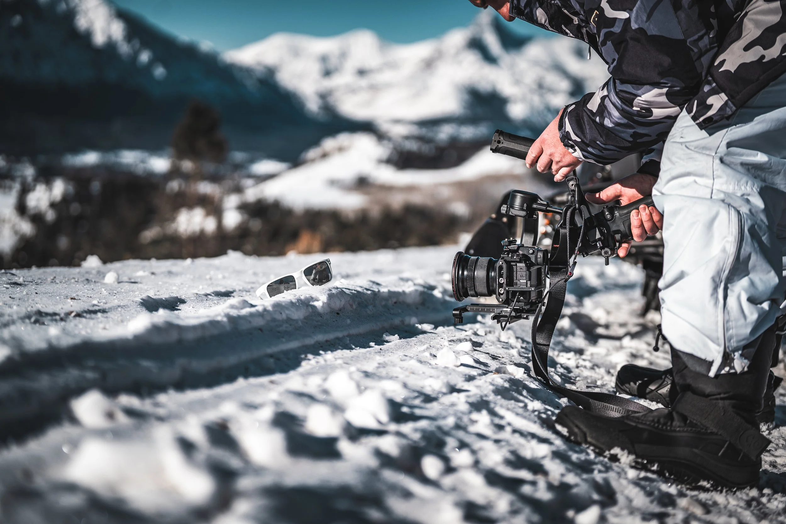 A person in winter clothing is kneeling in the snow with a professional video camera, recording a scene outdoors in a snowy mountain landscape.