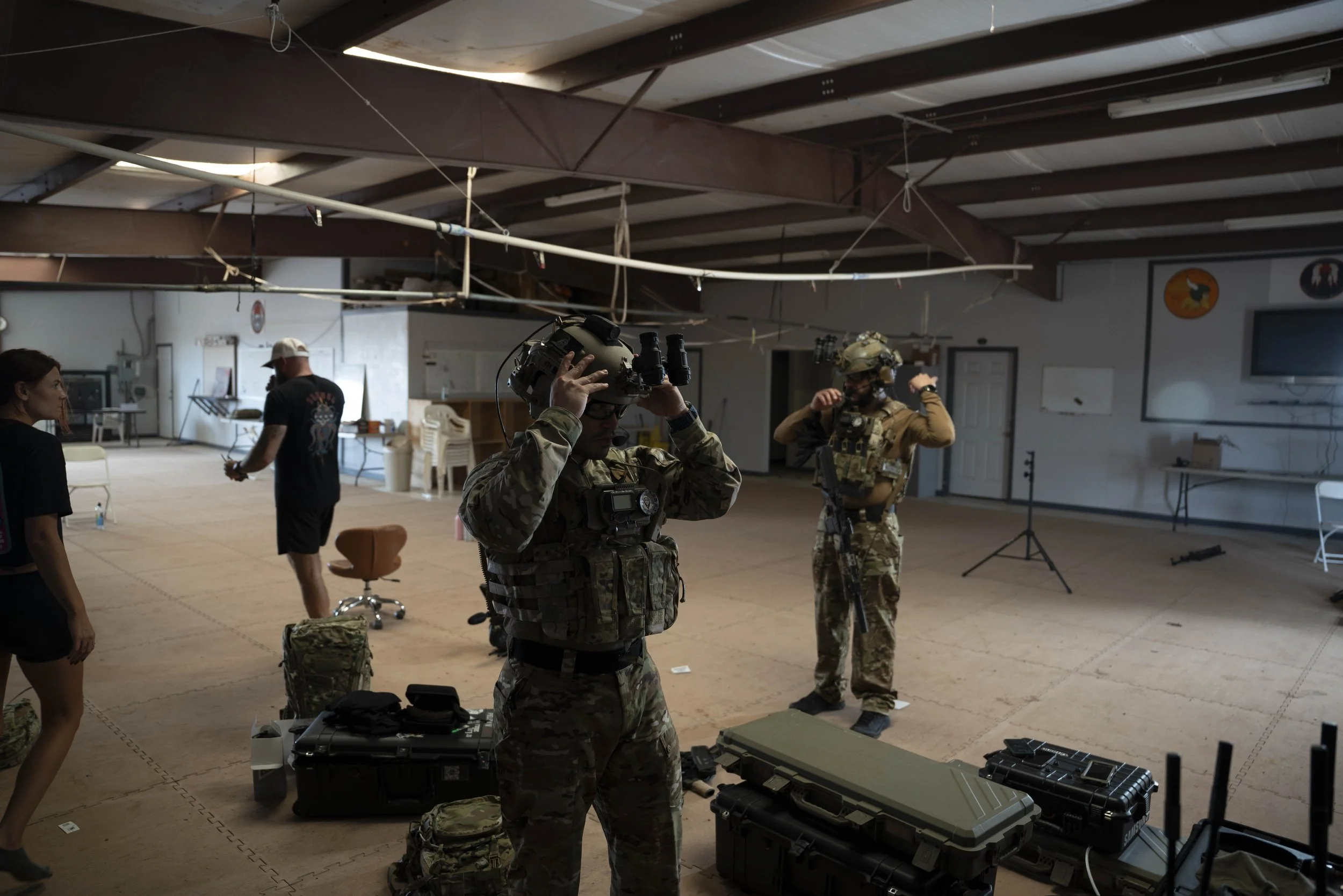 Two soldiers in military uniforms and tactical gear adjusting their helmets inside a spacious indoor training facility. There are various equipment and containers on the floor around them, with two civilians nearby and a TV screen on the wall.