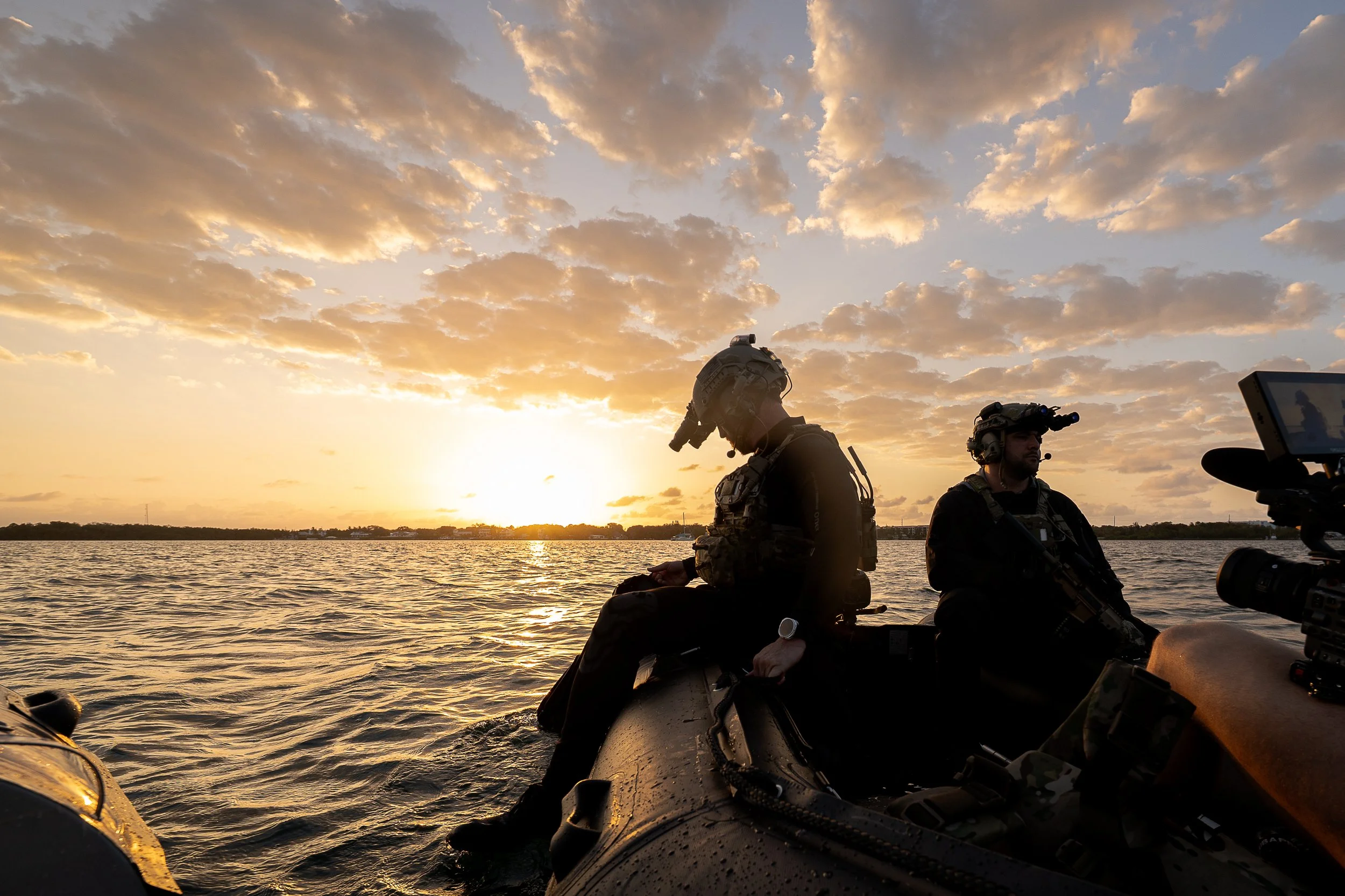 Two military personnel on a boat during sunset on the water, wearing tactical gear and helmets, with a camera filming nearby.