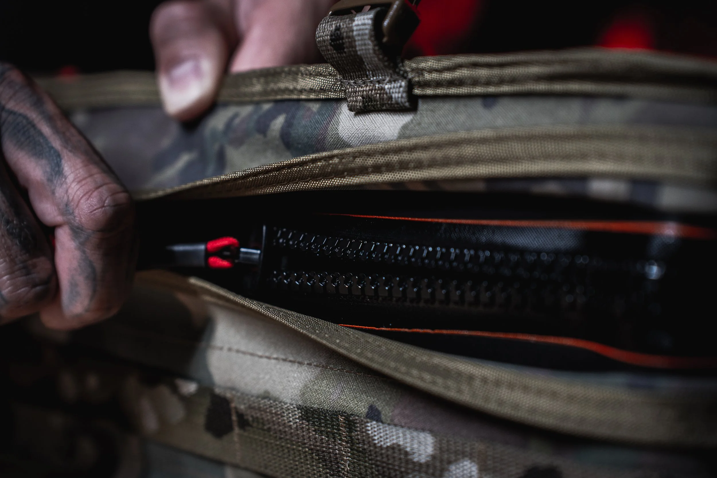 Close-up of a person's hand opening a tan camouflage tactical backpack, revealing a black zipper and some gear inside.