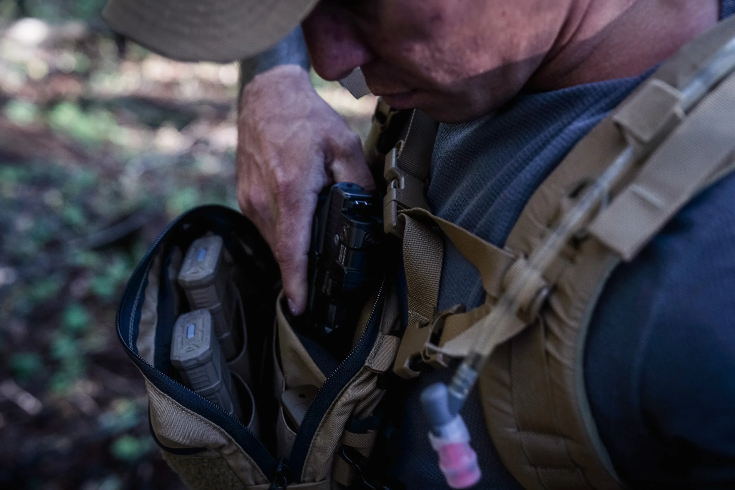 Close-up of a man in outdoor gear, retrieving a firearm from a tactical vest in a wooded environment.