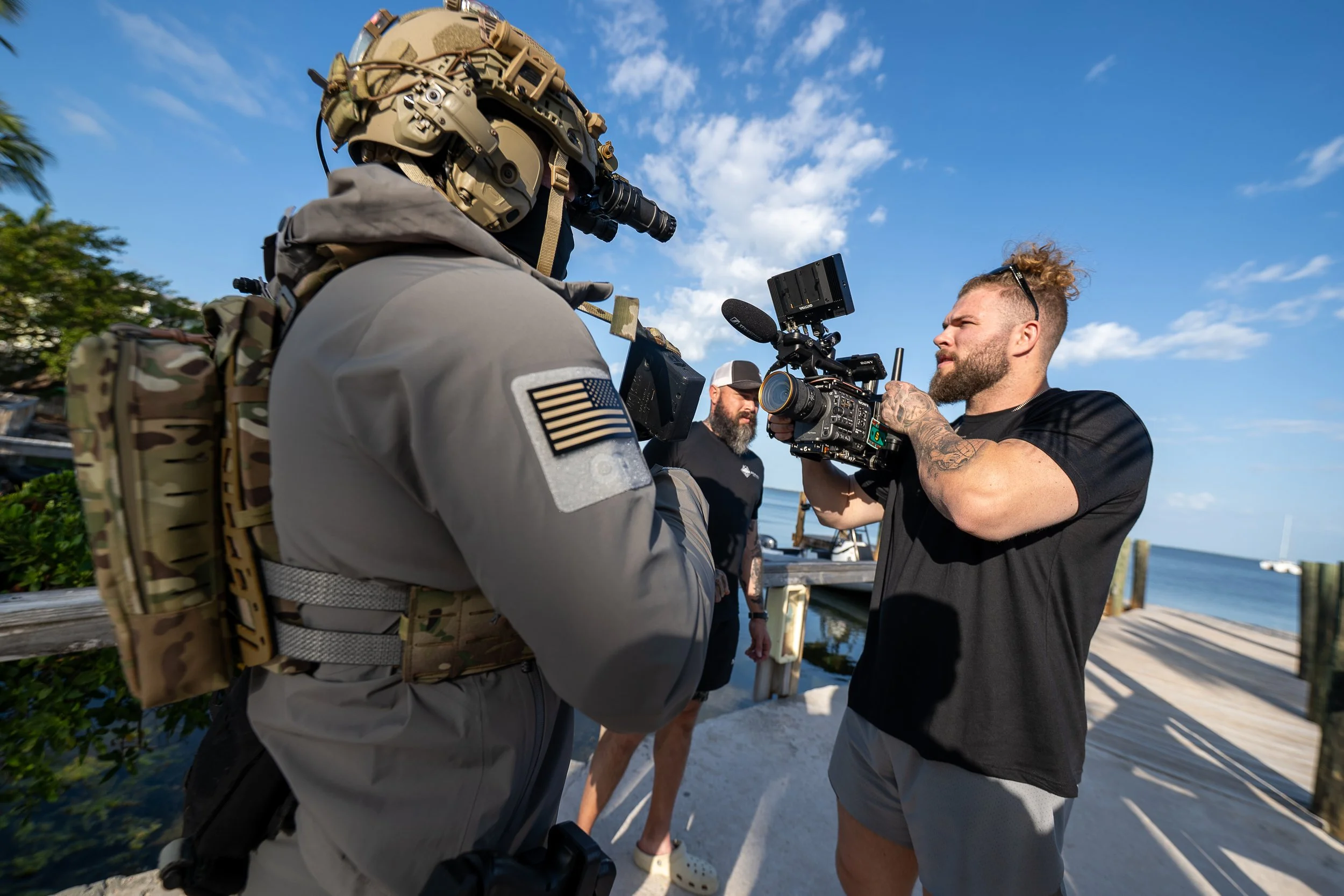 A man with a tattooed arm, beard, and sunglasses holding a camera is filming a person wearing military gear and a helmet with night vision equipment. Another man with a beard and black cap stands in the background on a pier near the ocean, under a bl