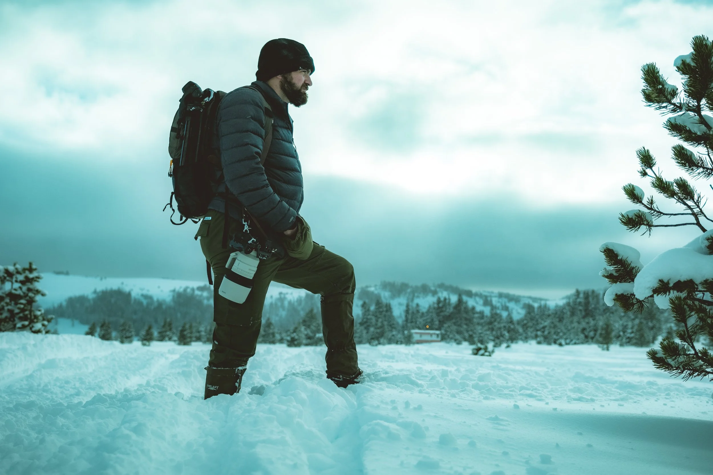 A man standing in snow-covered landscape with forests and cloudy sky, dressed in winter gear and carrying a backpack.