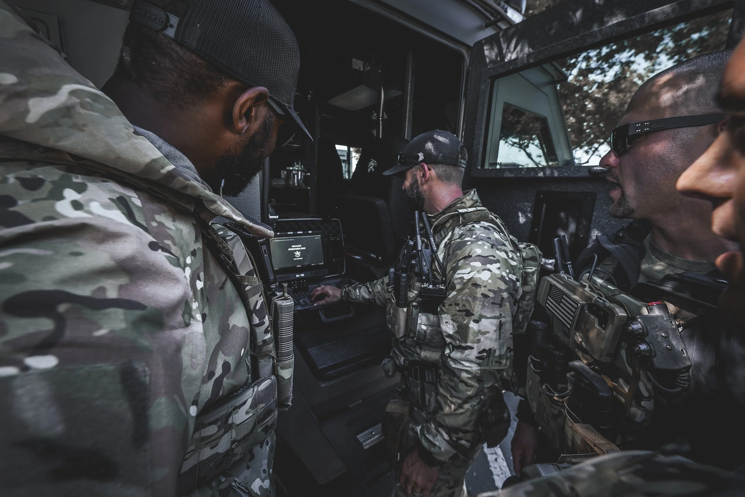 Four military personnel in camouflage uniforms and tactical gear inside a vehicle, gathered around a computer display, engaged in a discussion or briefing.