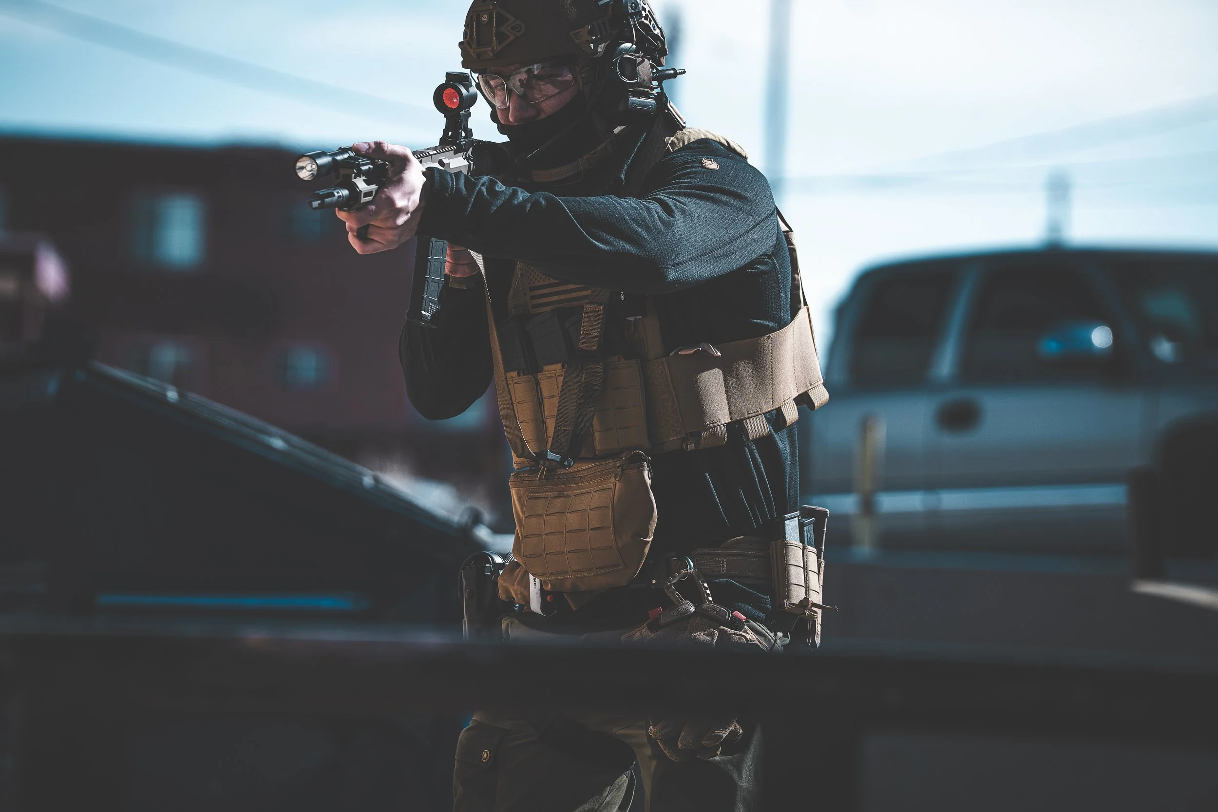 A soldier in combat gear aims a rifle with a red dot sight in an urban setting, wearing a helmet and tactical vest.