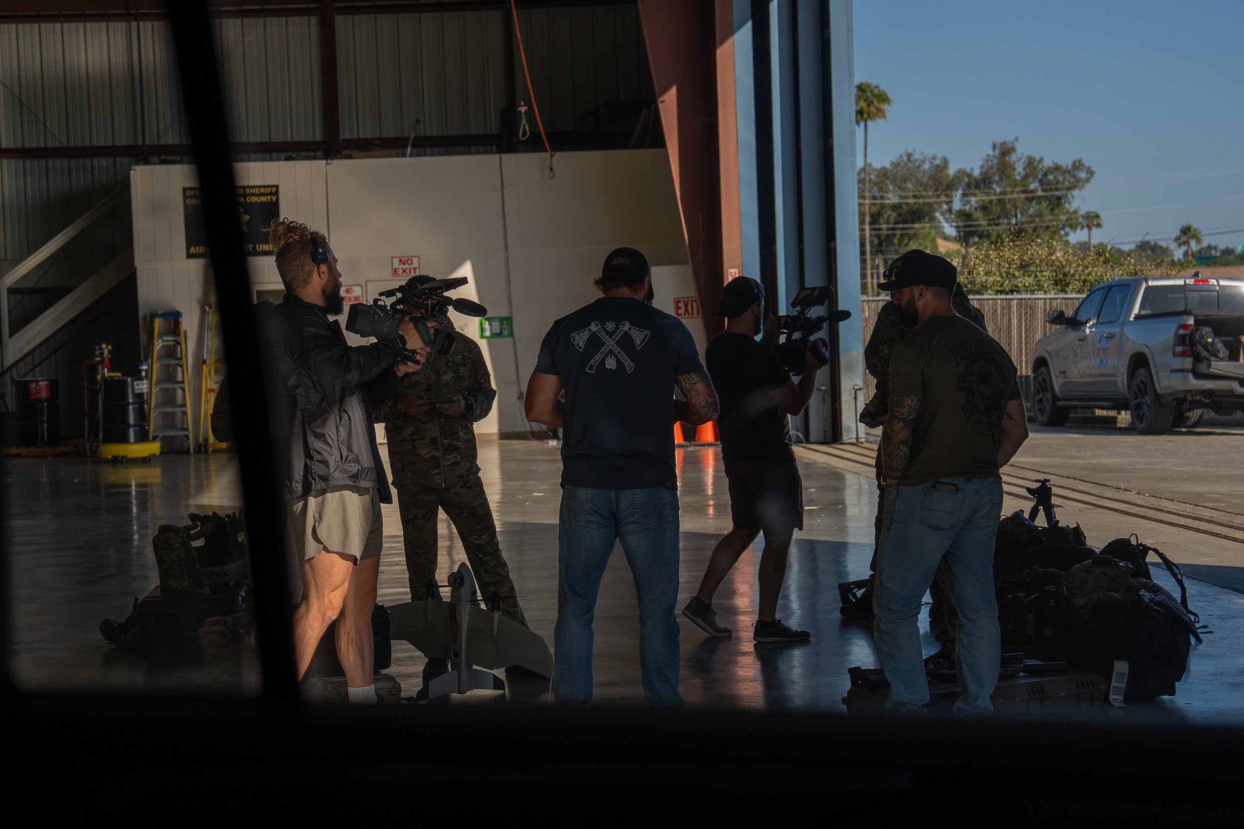 Group of people with camera equipment inside a spacious warehouse, some wearing military-style clothing, while others are dressed casually, with outside visible through the open doorway.
