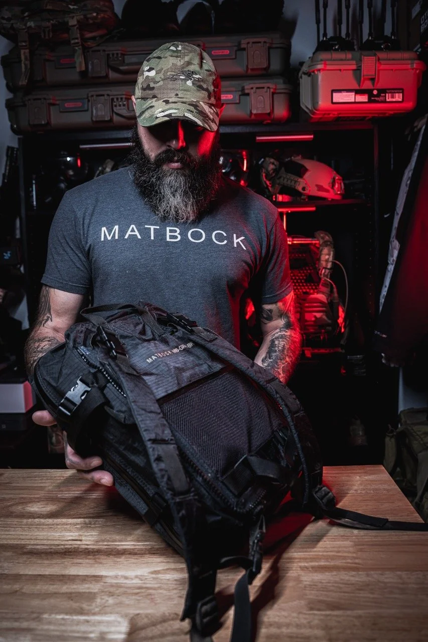 Man with a beard and camo cap holding a black tactical backpack inside a room with red ambient lighting and tactical gear on shelves.