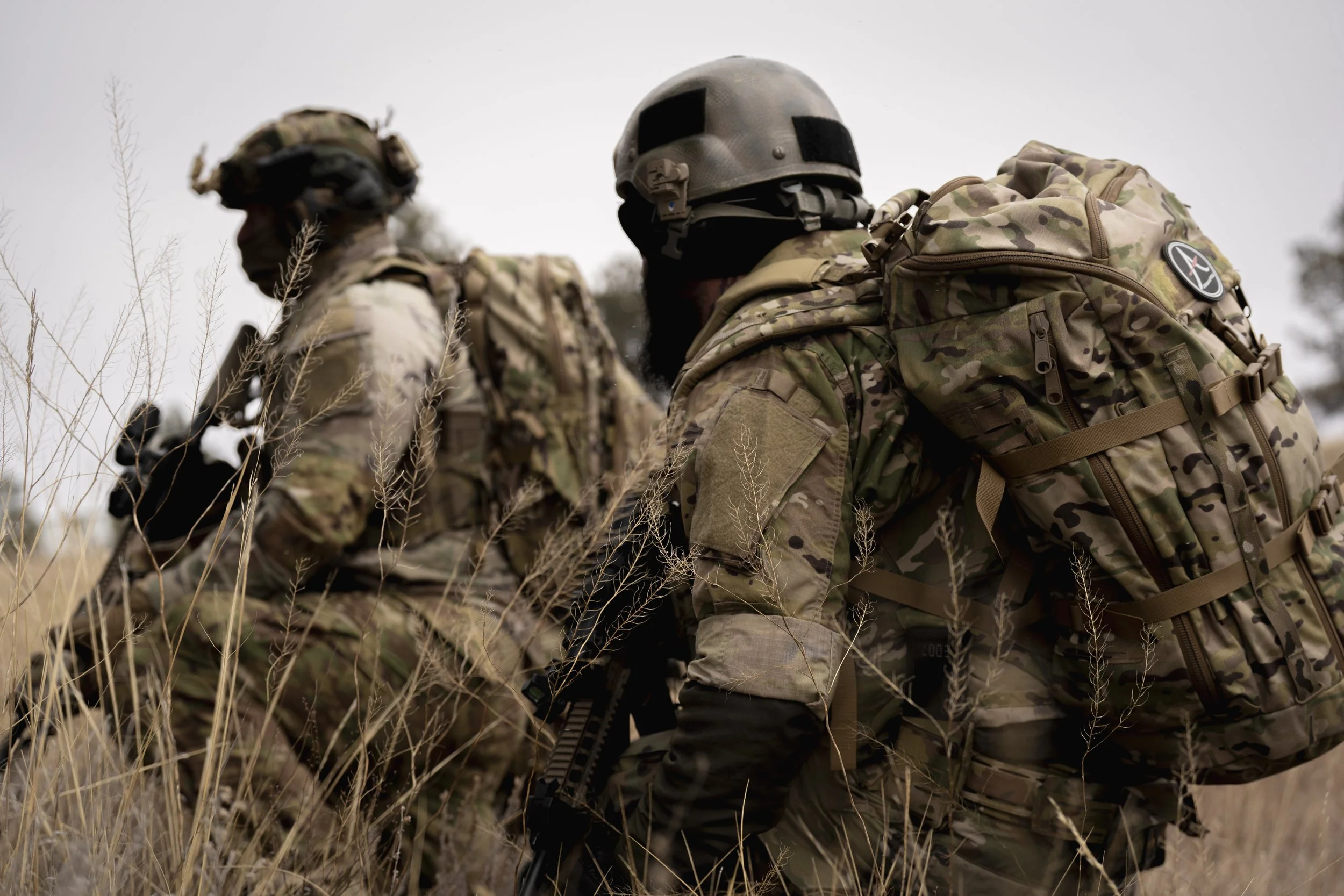 Two soldiers in camouflage uniforms with tactical gear and helmets, walking through tall grass in an outdoor setting.