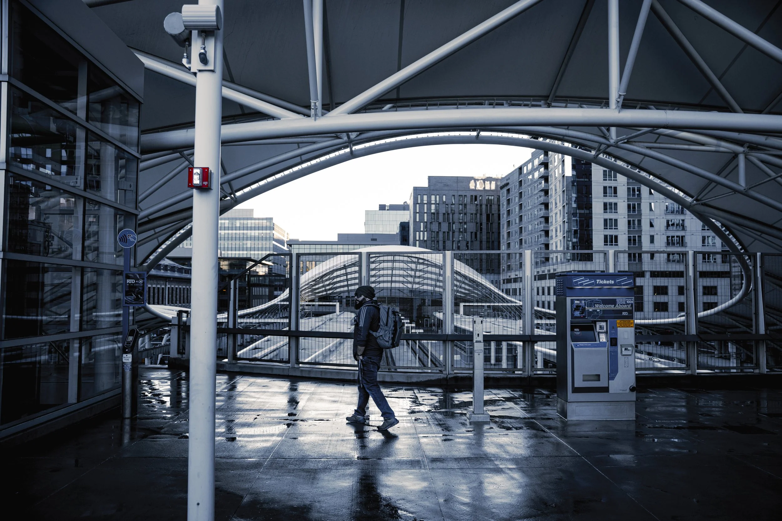 A person with a backpack walking through a modern urban train station with a covered platform, high rise buildings in the background, and ticket machines nearby.
