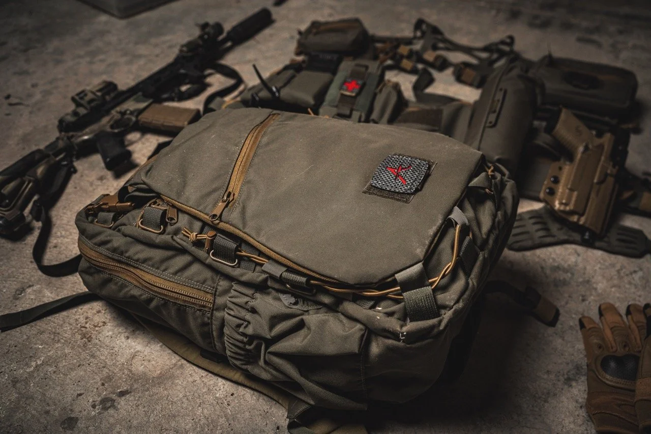 Military gear including a tactical backpack, rifles, a handgun, tactical gloves, and other combat equipment laid out on a concrete surface.