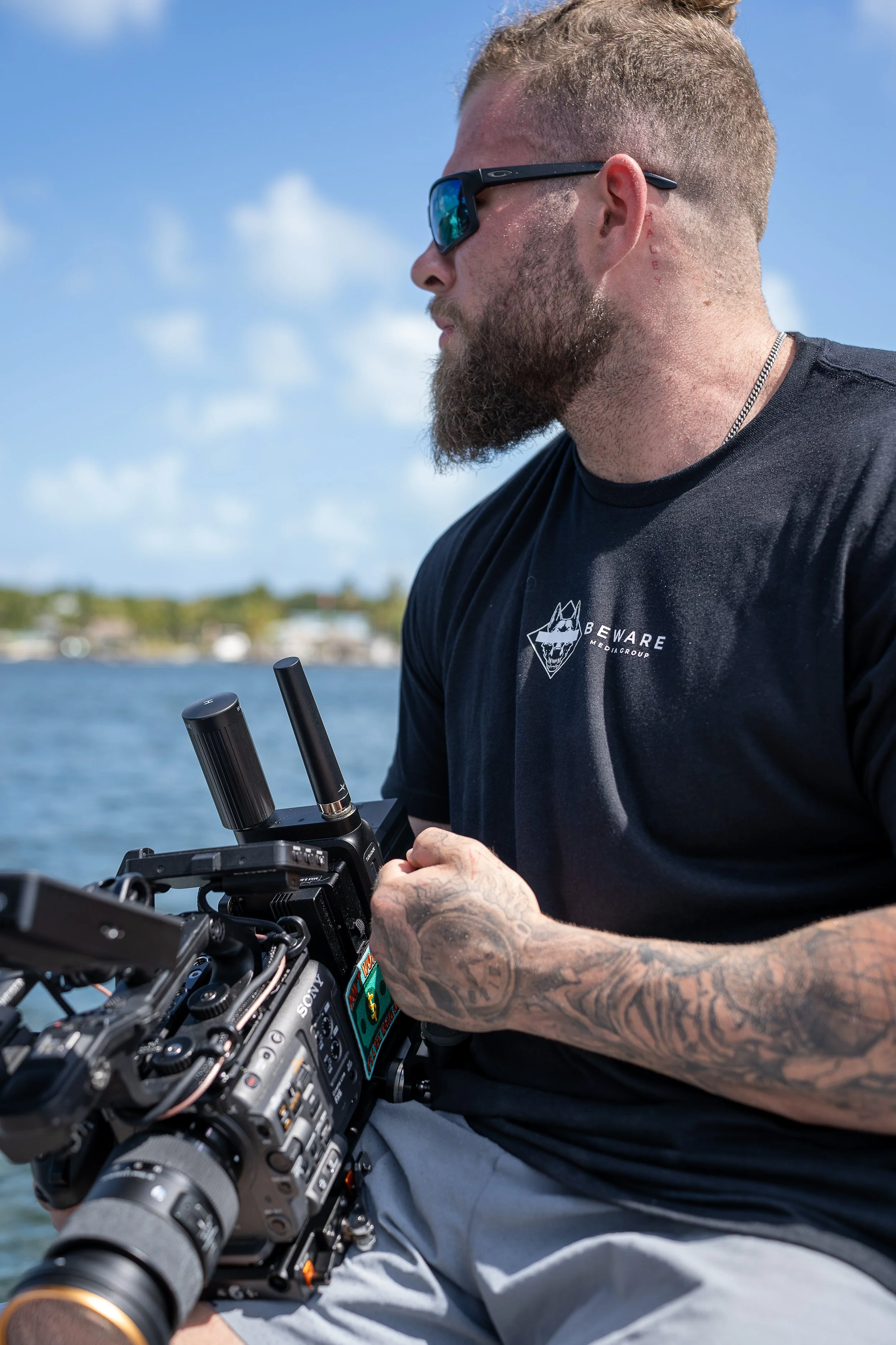 A man with tattoos, wearing sunglasses, a black T-shirt, and khaki shorts, is operating a professional video camera outdoors by a body of water with a shoreline and trees in the background.
