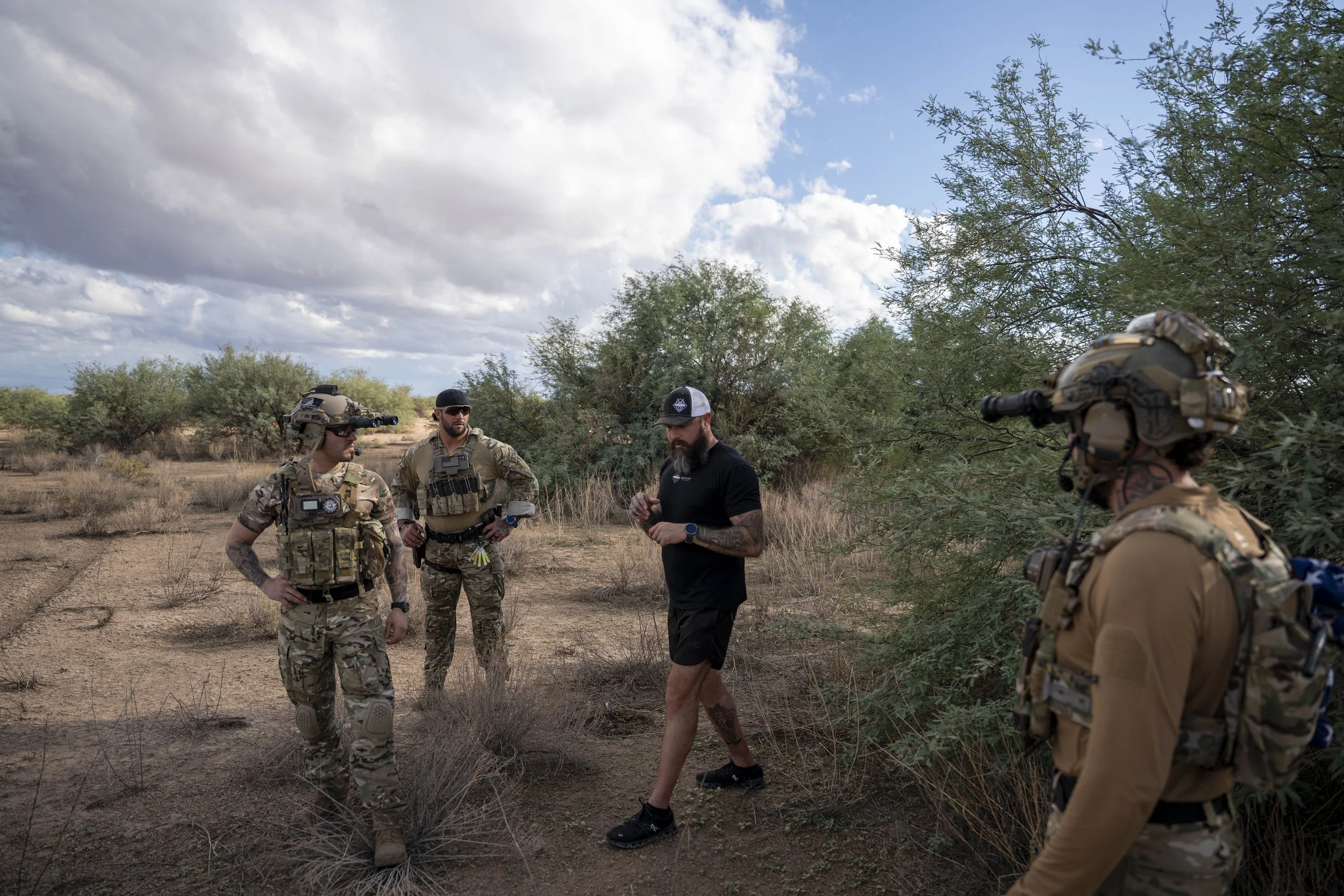 Four people outdoors in a desert landscape with sparse bushes, some wearing military gear and tactical equipment, while one is dressed casually in a black shirt and shorts. They appear to be engaged in a conversation.