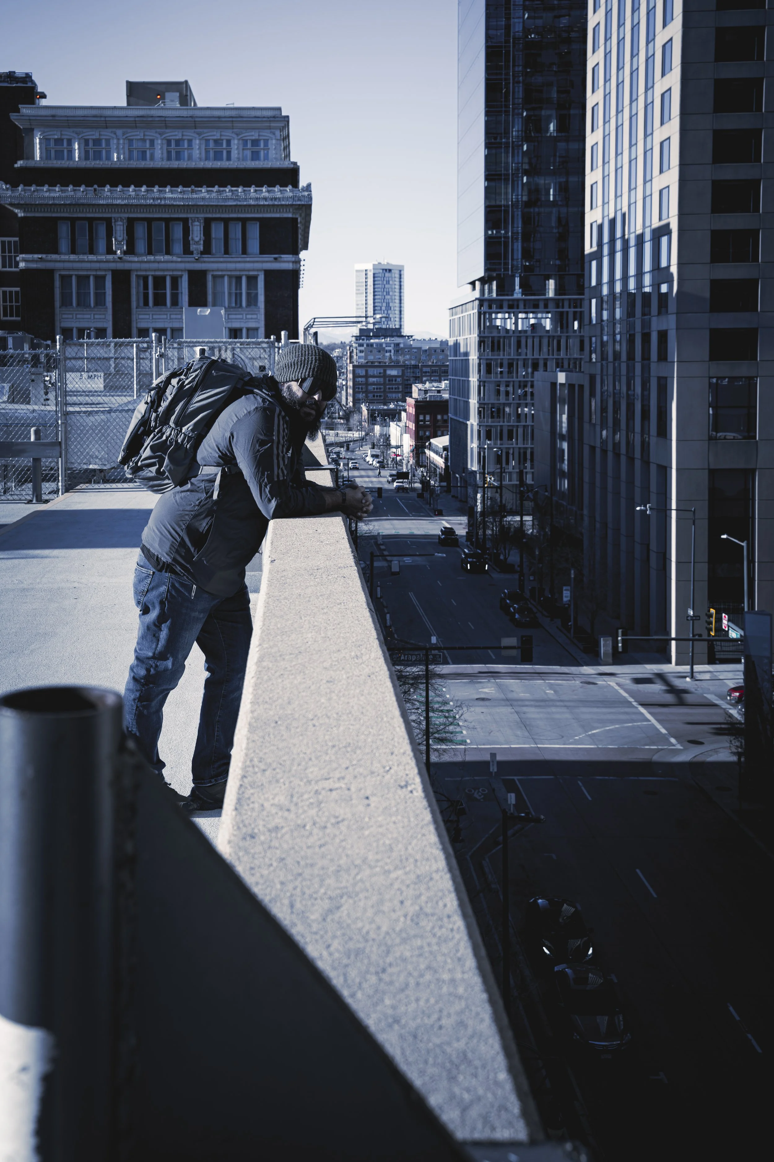 A man leaning on a concrete ledge on a high-rise rooftop, overlooking a city street with tall buildings, cars, and sparse traffic during daytime.