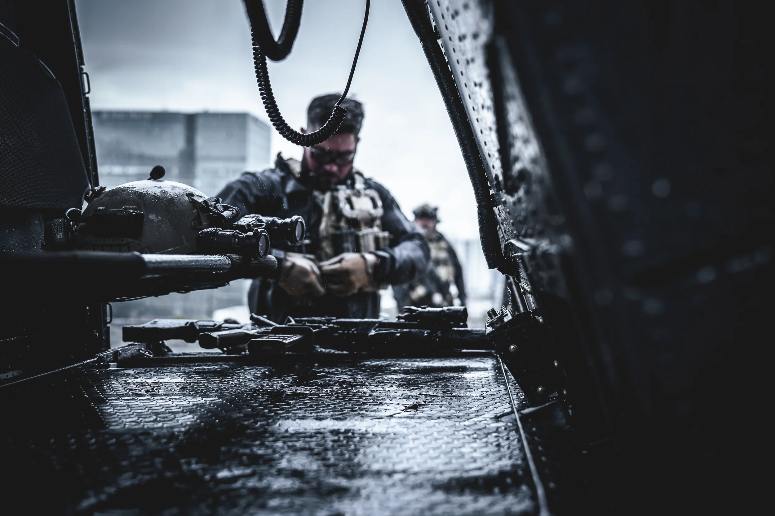 Two soldiers in tactical gear loading weapons inside a military vehicle with rain on the surface.
