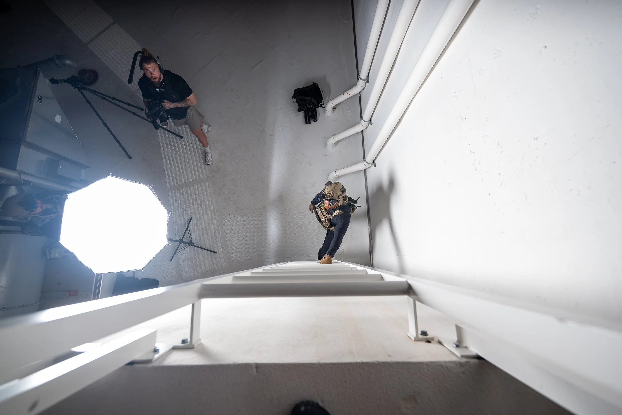 A photo taken from the top of a ladder showing a man in tactical gear and another person with camera equipment in a room with white walls and pipes, illuminated by a large softbox light.