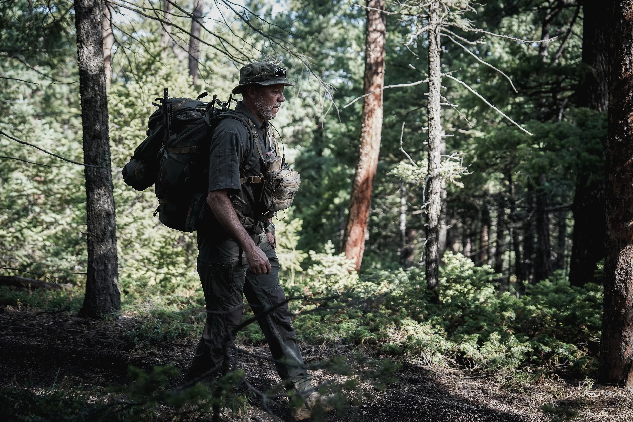 An older man hiking through a dense forest with tall trees, wearing outdoor clothing, a hat, and carrying a large backpack.