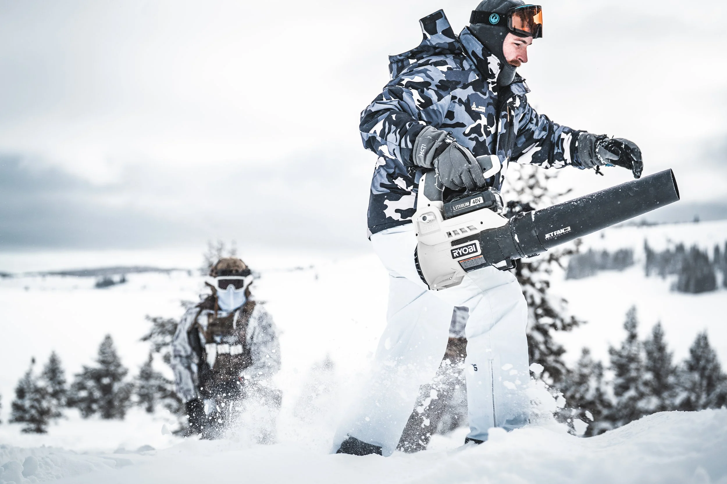 Two men in winter gear using a leaf blower in a snowy outdoor landscape.