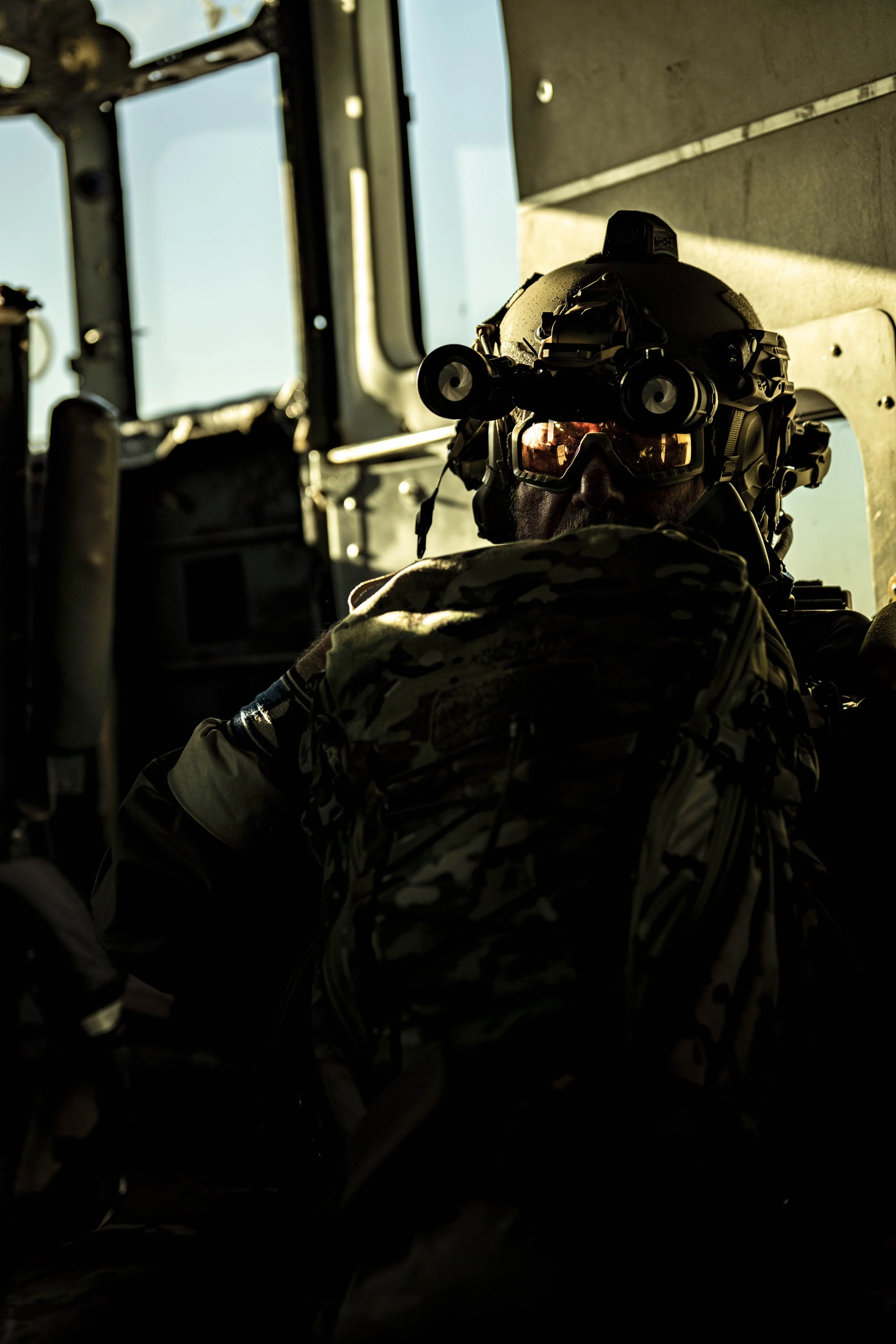 A soldier wearing camouflage uniform, tactical helmet with night vision goggles, and sunglasses, sitting inside a military vehicle in front of a window.