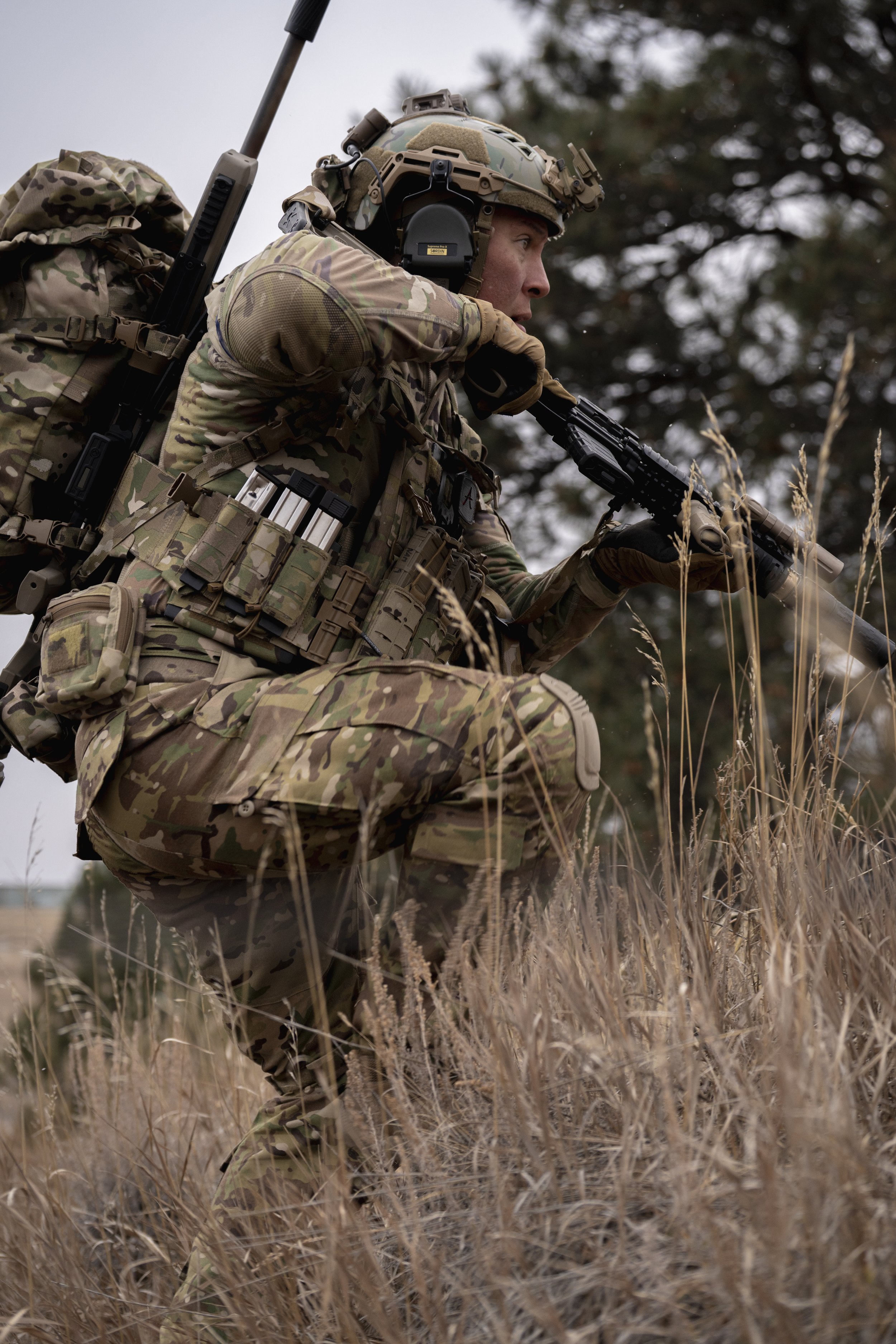 A soldier in camouflage uniform kneeling in tall grass while aiming a rifle outdoors.