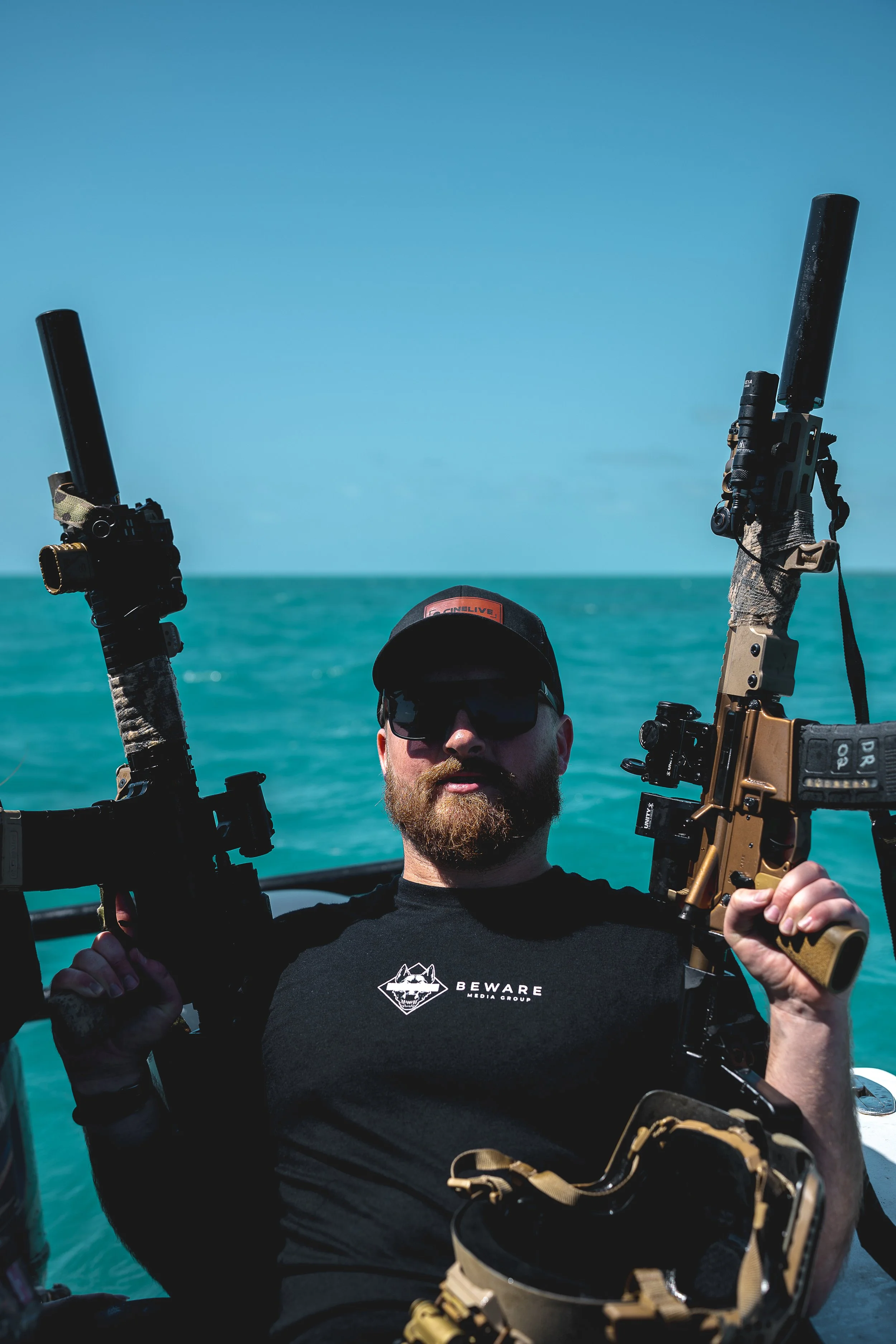 Man with a beard wearing sunglasses and a black cap, holding two rifles, aboard a boat in the ocean with a clear blue sky.