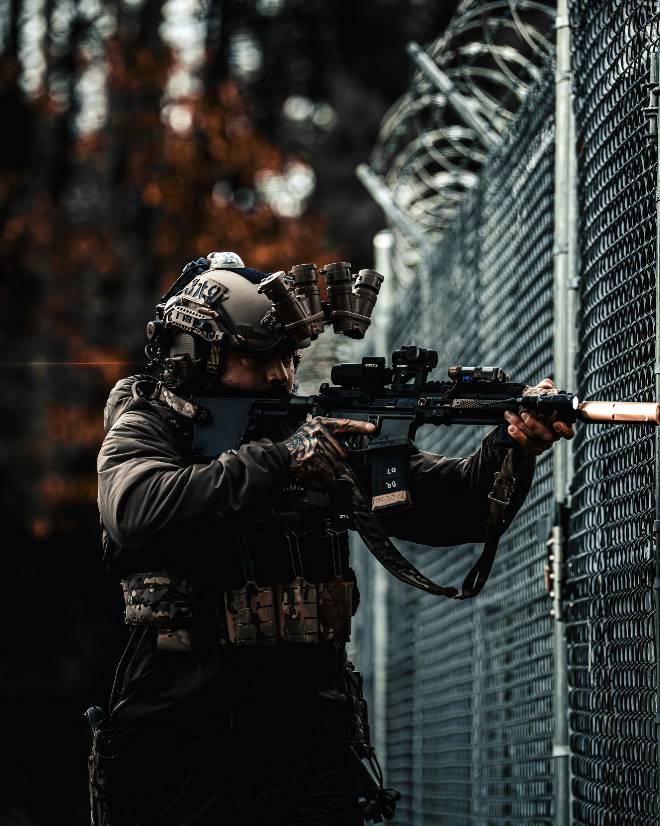 A soldier wearing tactical gear and night-vision goggles fires an assault rifle through a fence.