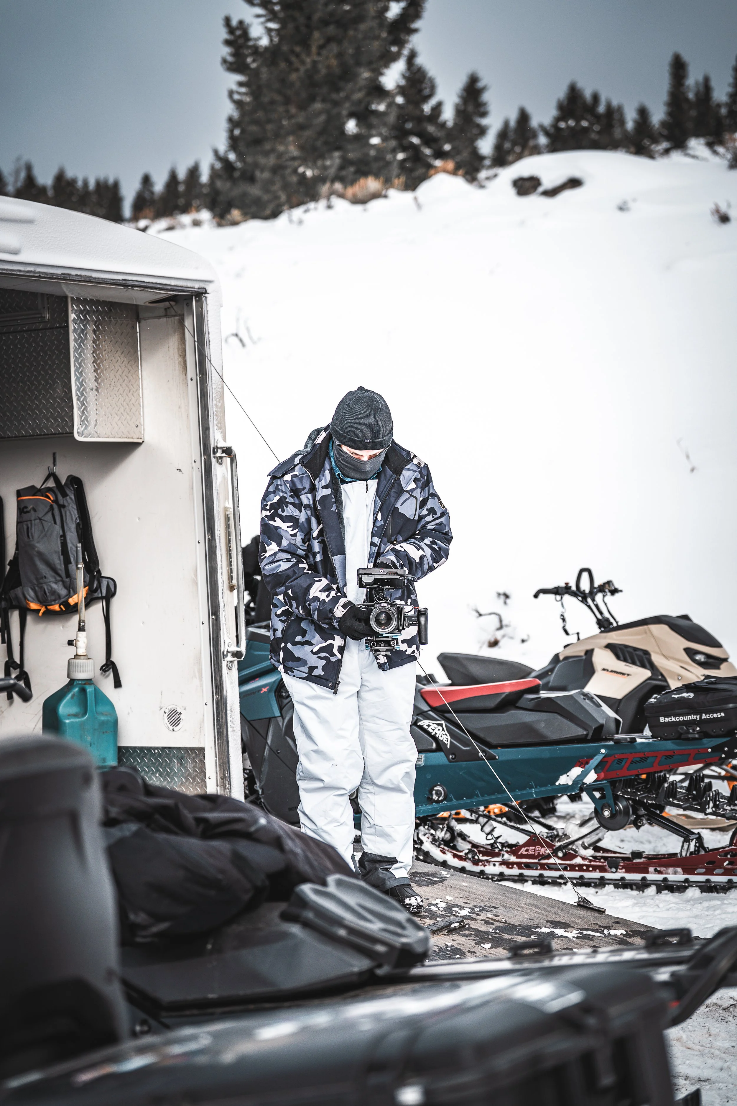 A person dressed in winter gear with a face mask holding a camera, standing beside a snowmobile and a trailer with snow-covered ground and trees in the background.