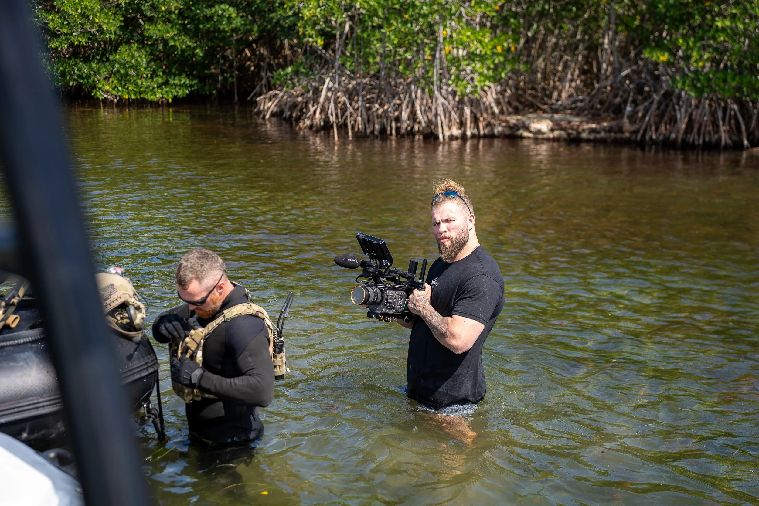 Two men standing in water near a shoreline with mangroves, one holding a professional camera and the other adjusting gear on a boat.