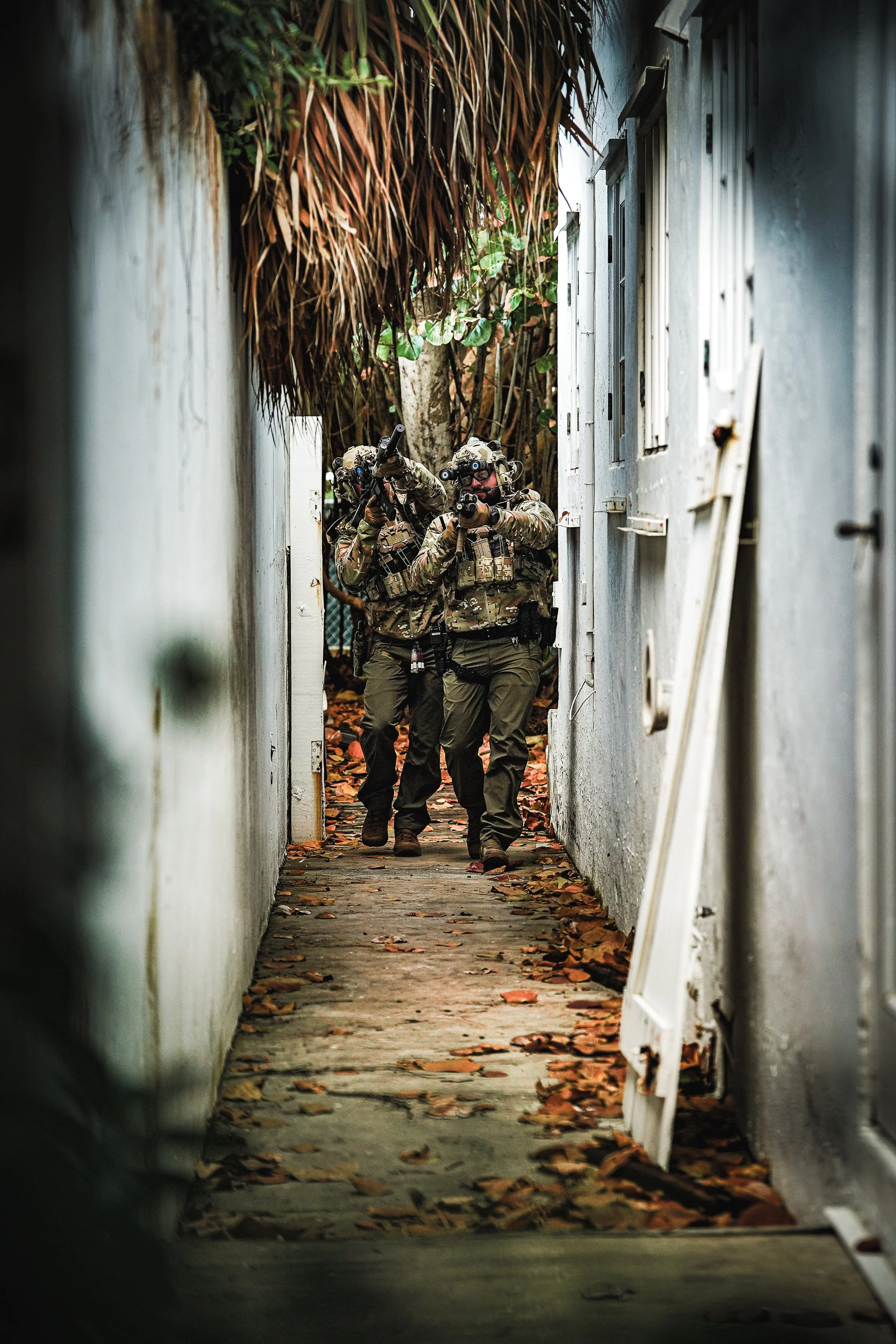 Two soldiers in combat gear walking through a narrow alleyway with fallen leaves on the ground, flanked by white walls and a doorway on the right.