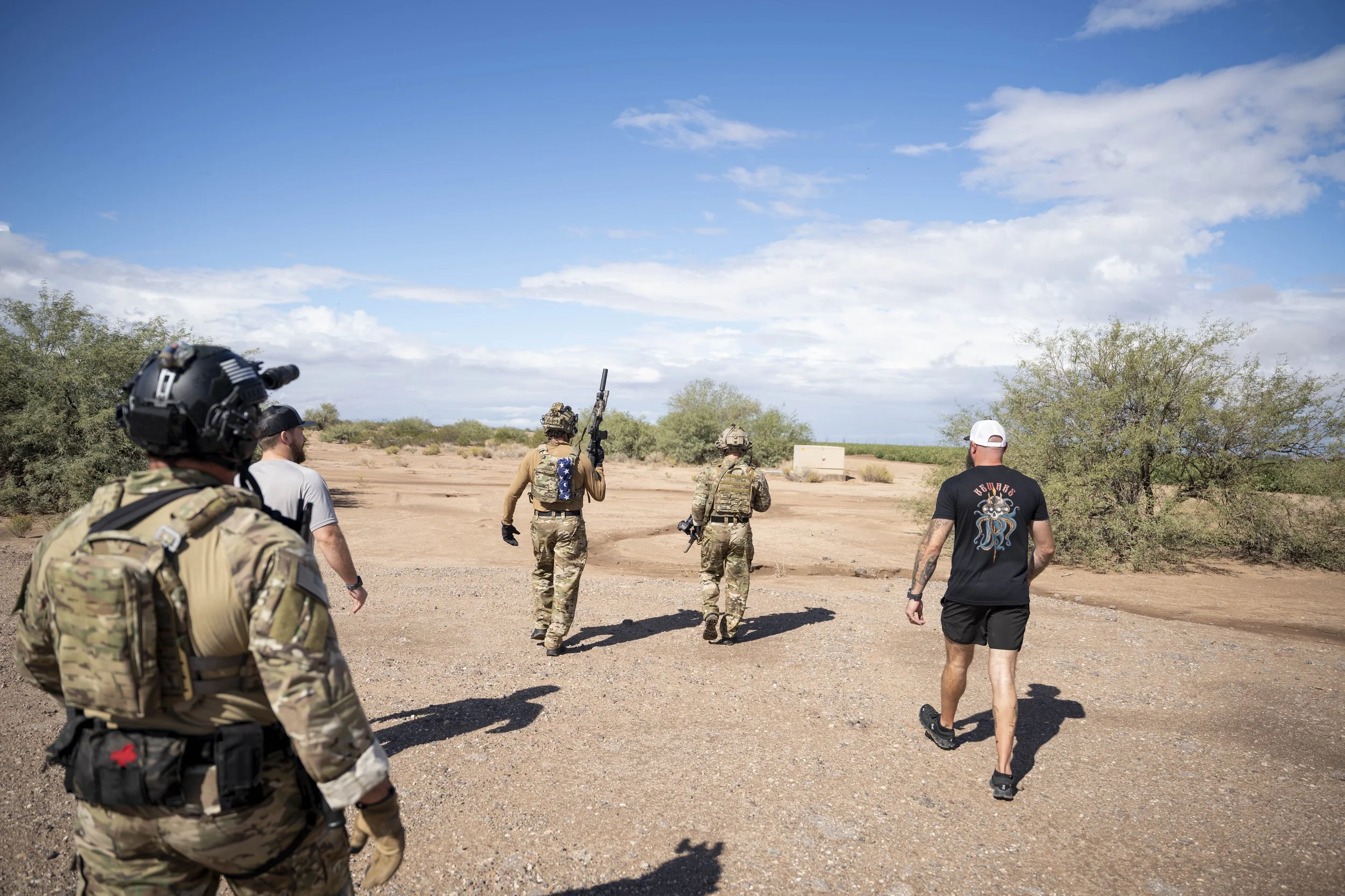 Four military personnel in camouflage uniforms and two civilians walking in a desert landscape with sparse vegetation under a partly cloudy sky.
