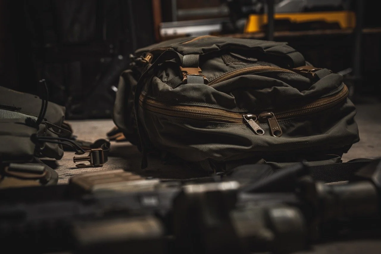 A black backpack with brown zippers placed on a dark surface, with other gear and equipment nearby in a dimly lit setting.