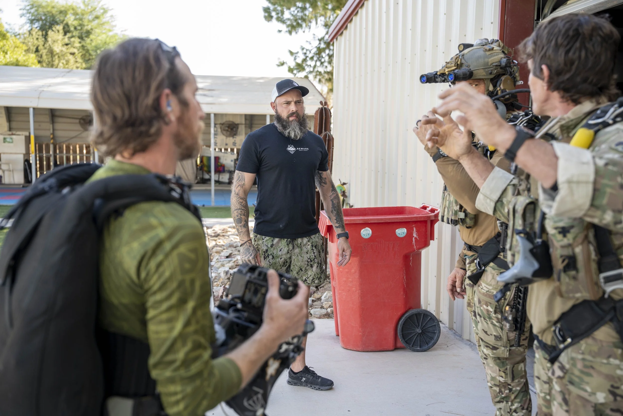 Group of people, likely soldiers or military personnel, standing outdoors and listening to a man with a beard, tattoos, wearing a black T-shirt and camouflage shorts. The group includes individuals with tactical gear and a man with night vision goggl