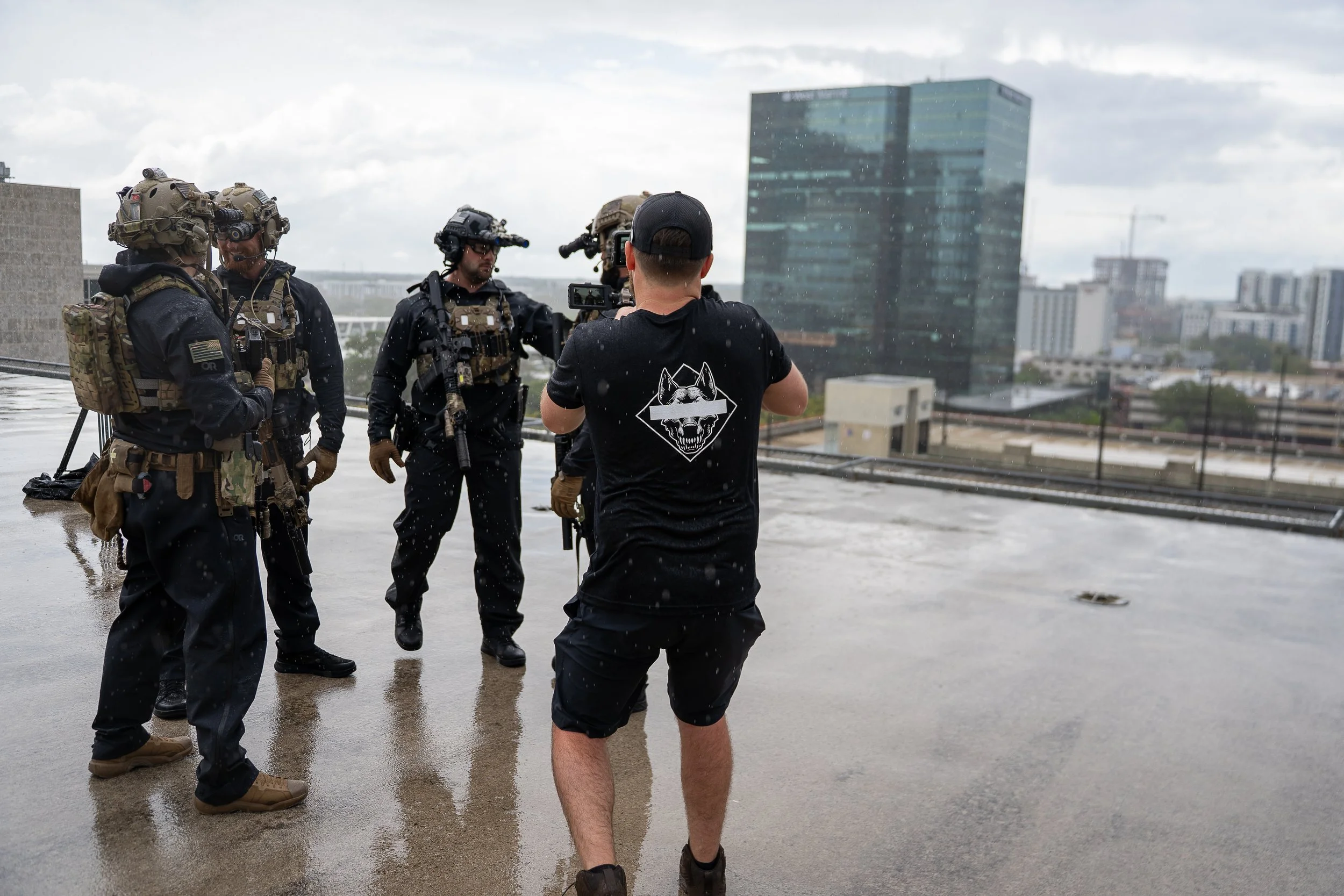 A man taking a photo of a group of four armed military or tactical personnel on a rooftop during rainy weather, with a city skyline in the background.