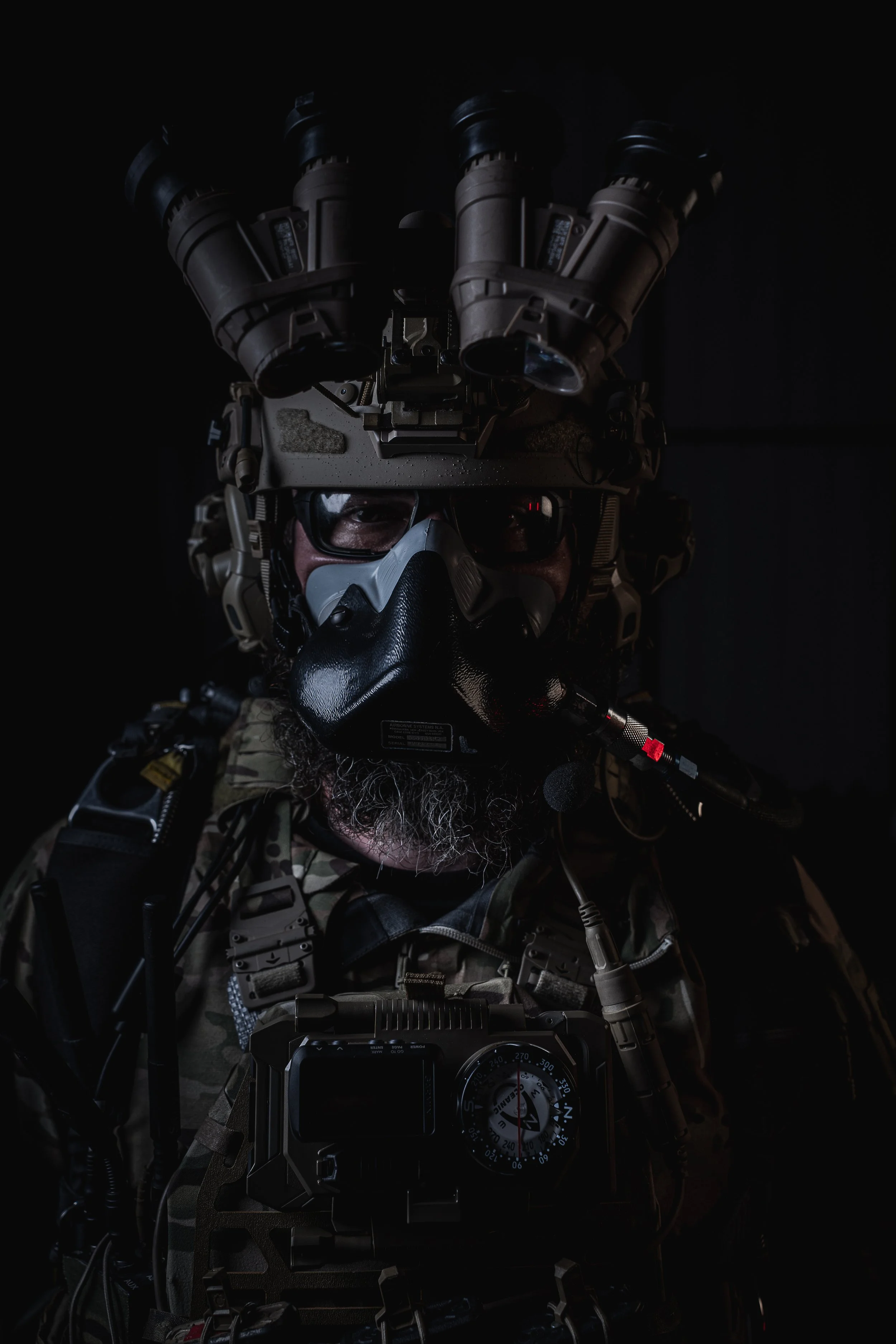 A soldier wearing tactical gear, camouflage uniform, and specialized equipment including night-vision goggles, a mask, and a helmet with mounted devices, against a dark background.