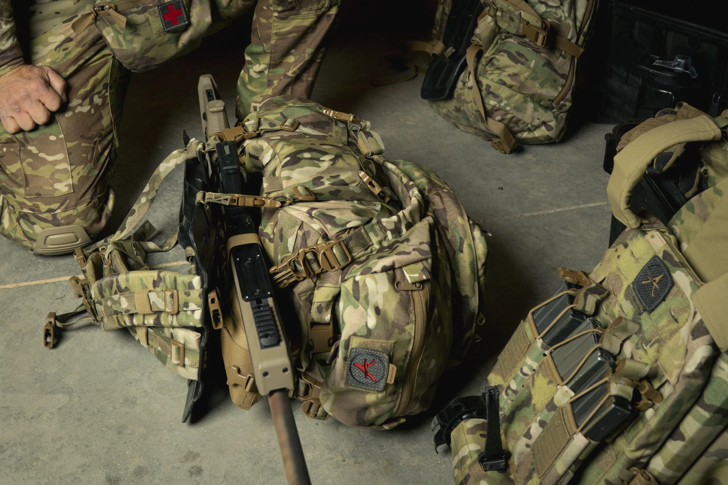 Military camouflage backpacks and tactical gear on the floor, with a person in camouflage uniform nearby.