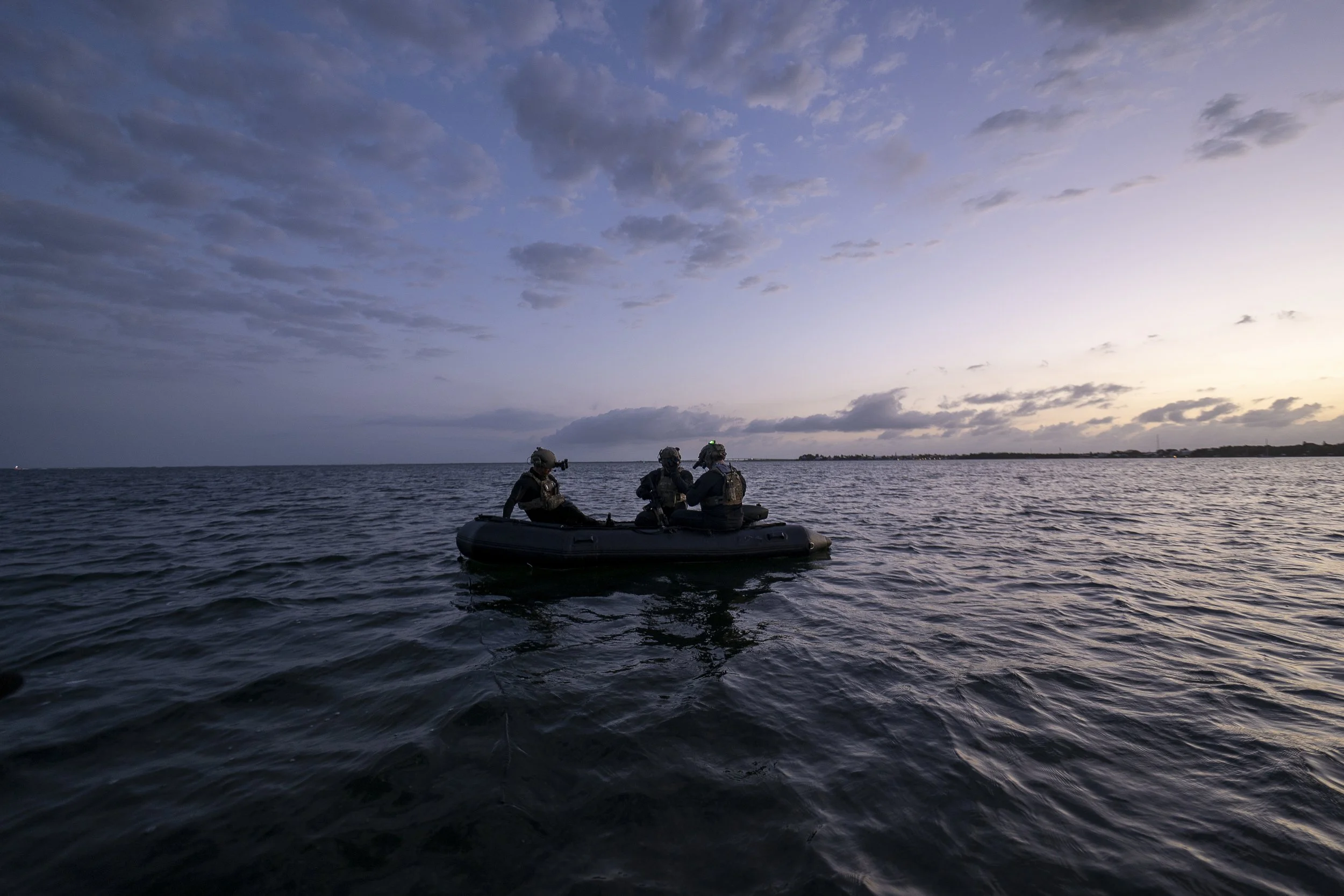 Three people in uniform on a small inflatable boat on the water at sunset, with a cloudy sky.