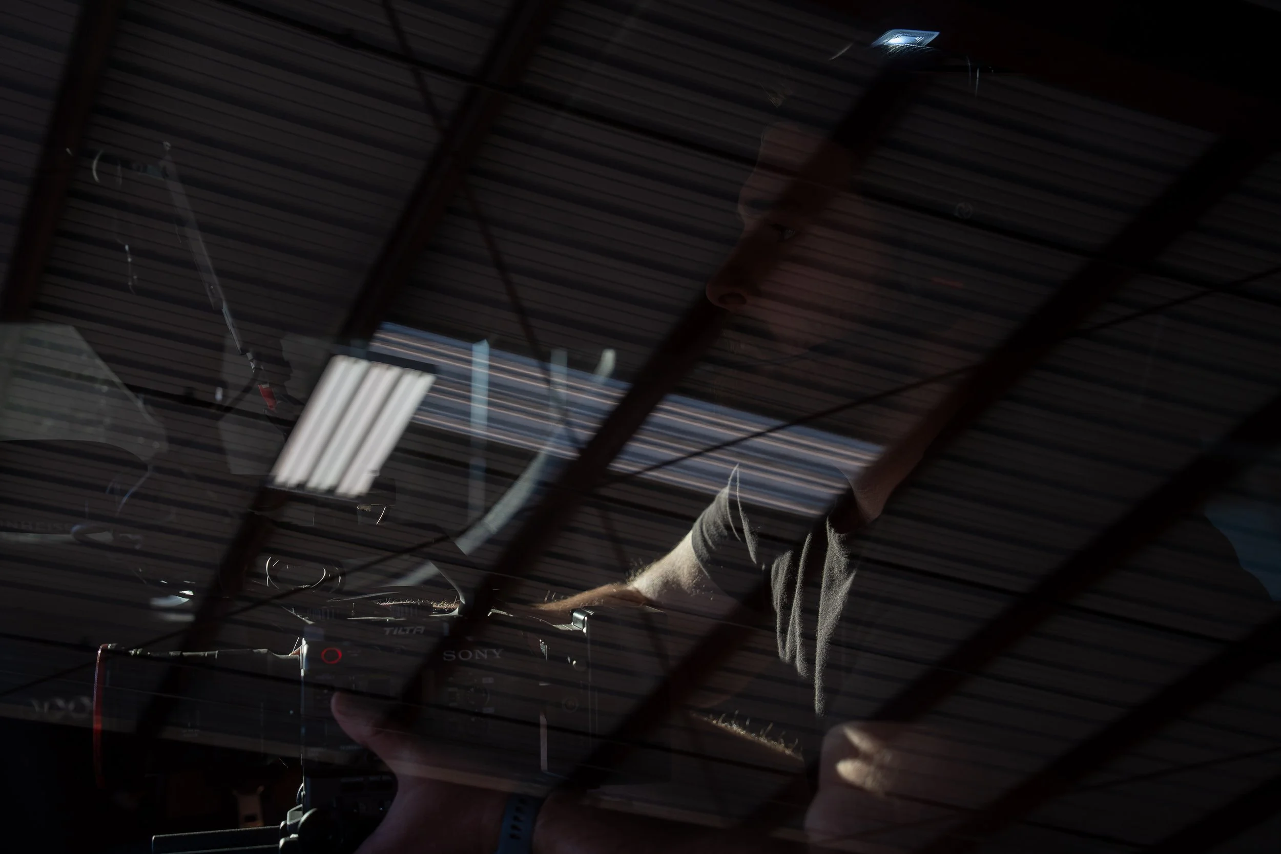 A person is working at a computer or DJ equipment, with their reflection visible on a glass surface that has a metal grid pattern.