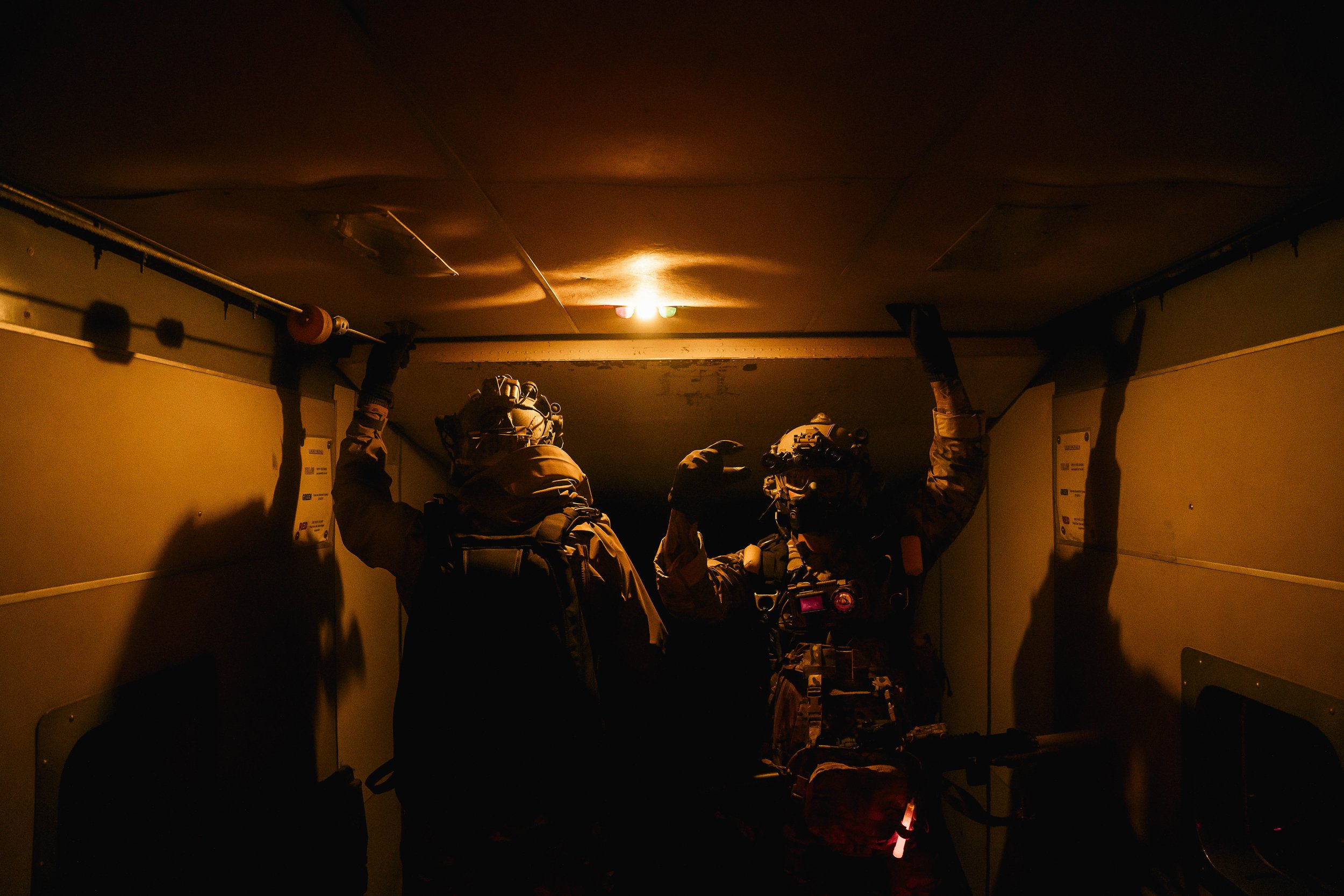 Two soldiers in tactical gear standing in a dimly lit corridor, silhouetted by a small ceiling light, with backpacks and helmets.