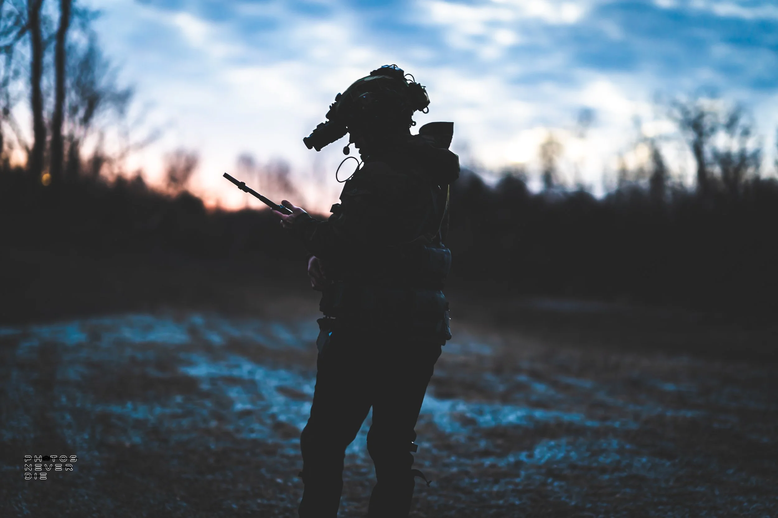 Silhouette of a soldier standing outdoors during dusk or dawn, looking at a handheld device, with bare trees in the background.