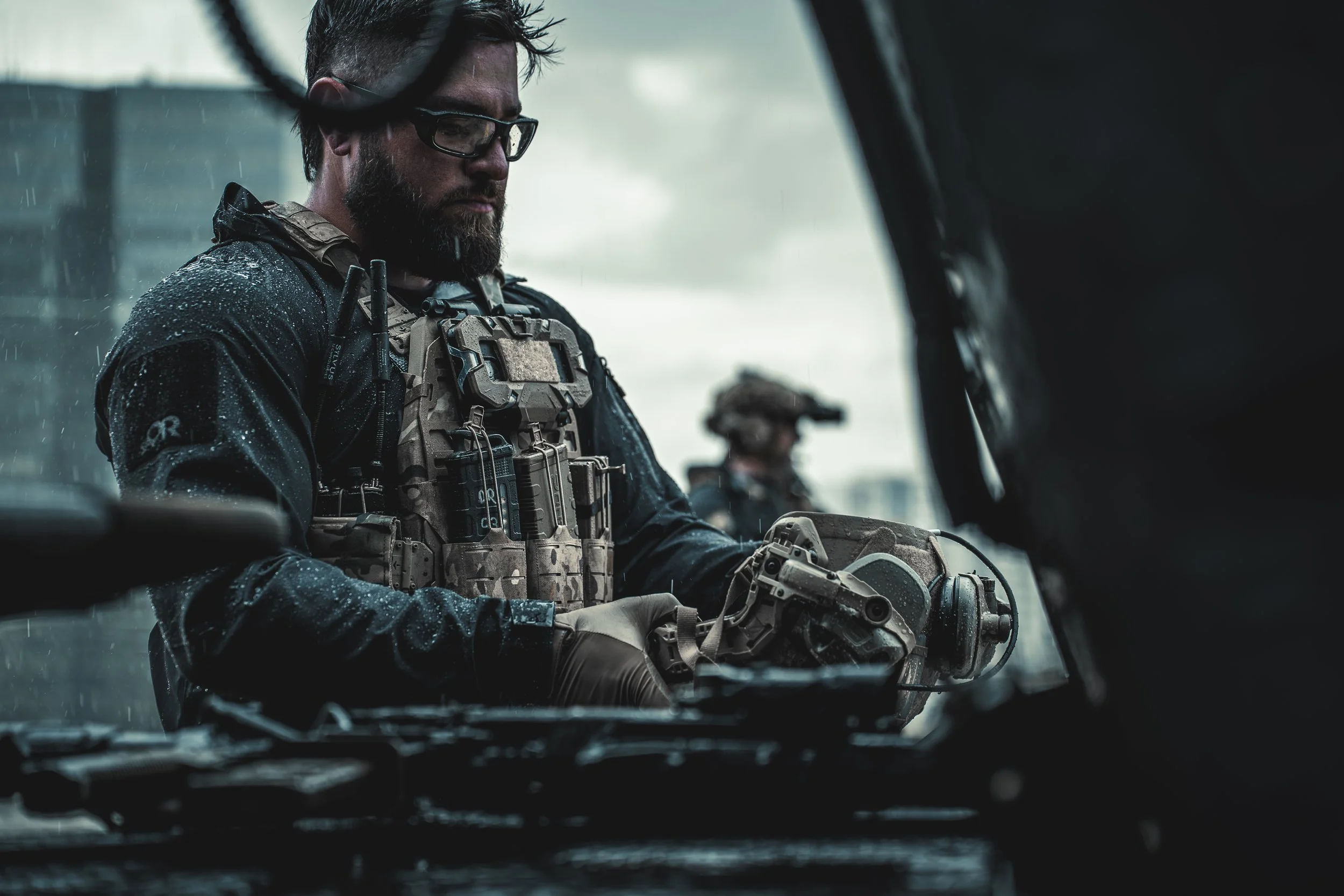 A man wearing tactical gear, gloves, and glasses inspects a firearm inside a vehicle during rainy weather, with another person in similar gear visible in the background.