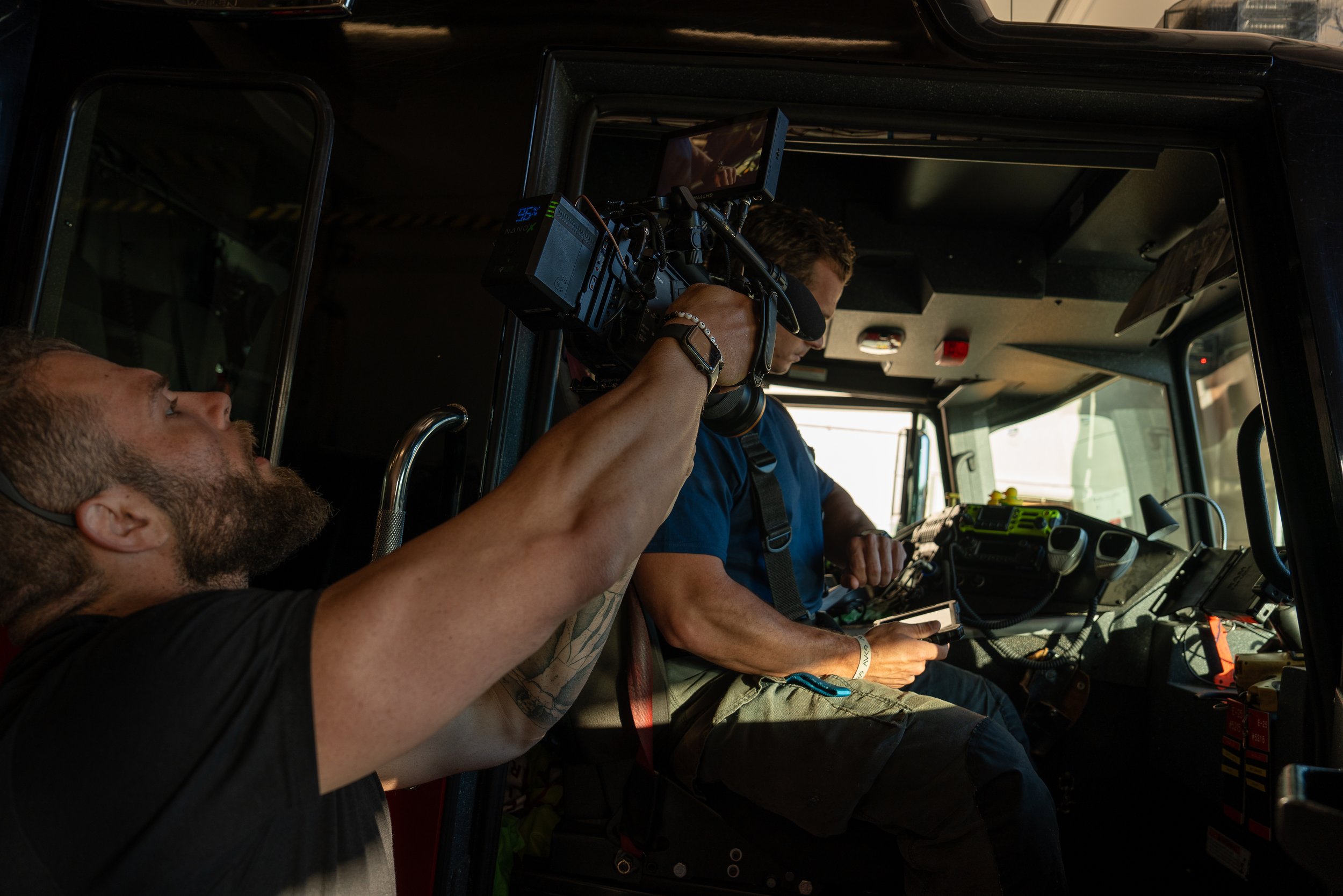 Two men inside a vehicle, one is operating a camera mounted on his shoulder, and the other is looking at a device in his hand. The scene appears to be on a film or video shoot.