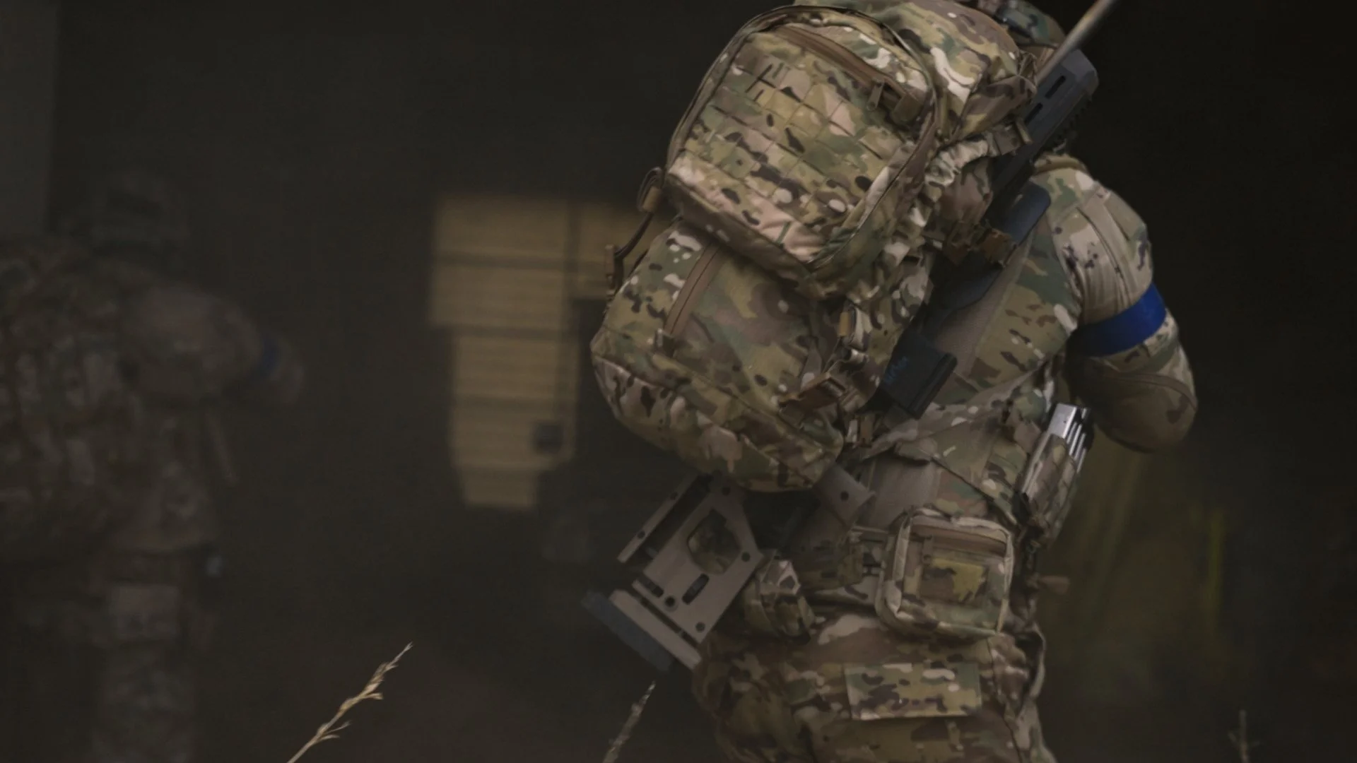 A soldier dressed in camouflage military gear, carrying a backpack and a firearm, walking in a dark environment.