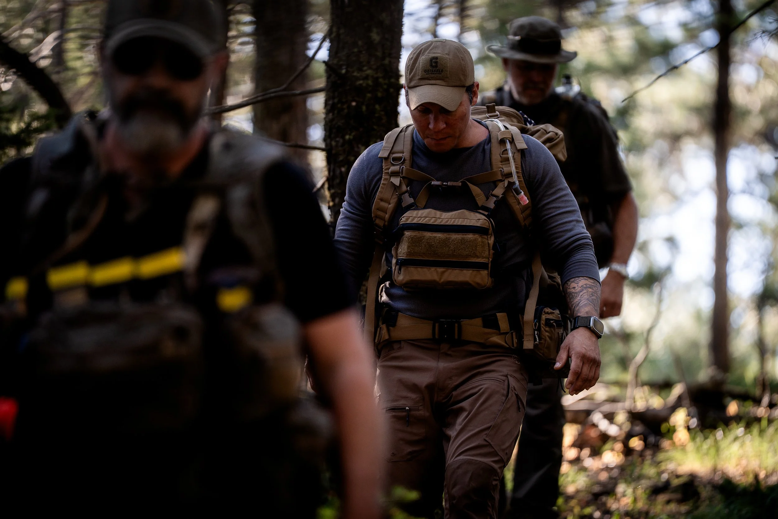 Three men hiking through a wooded forest with backpacks, wearing outdoor gear and hats, surrounded by trees and sunlight.