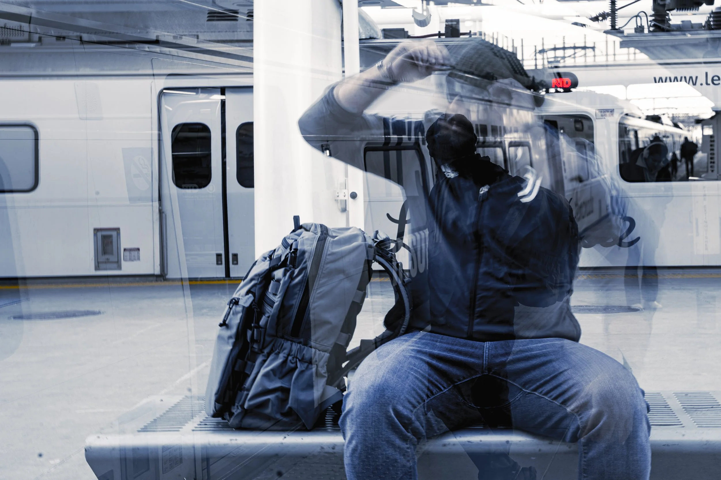 A person sitting on a bench at an airport, wearing a jacket and cap, with a backpack beside them, seen through a glass window or wall.