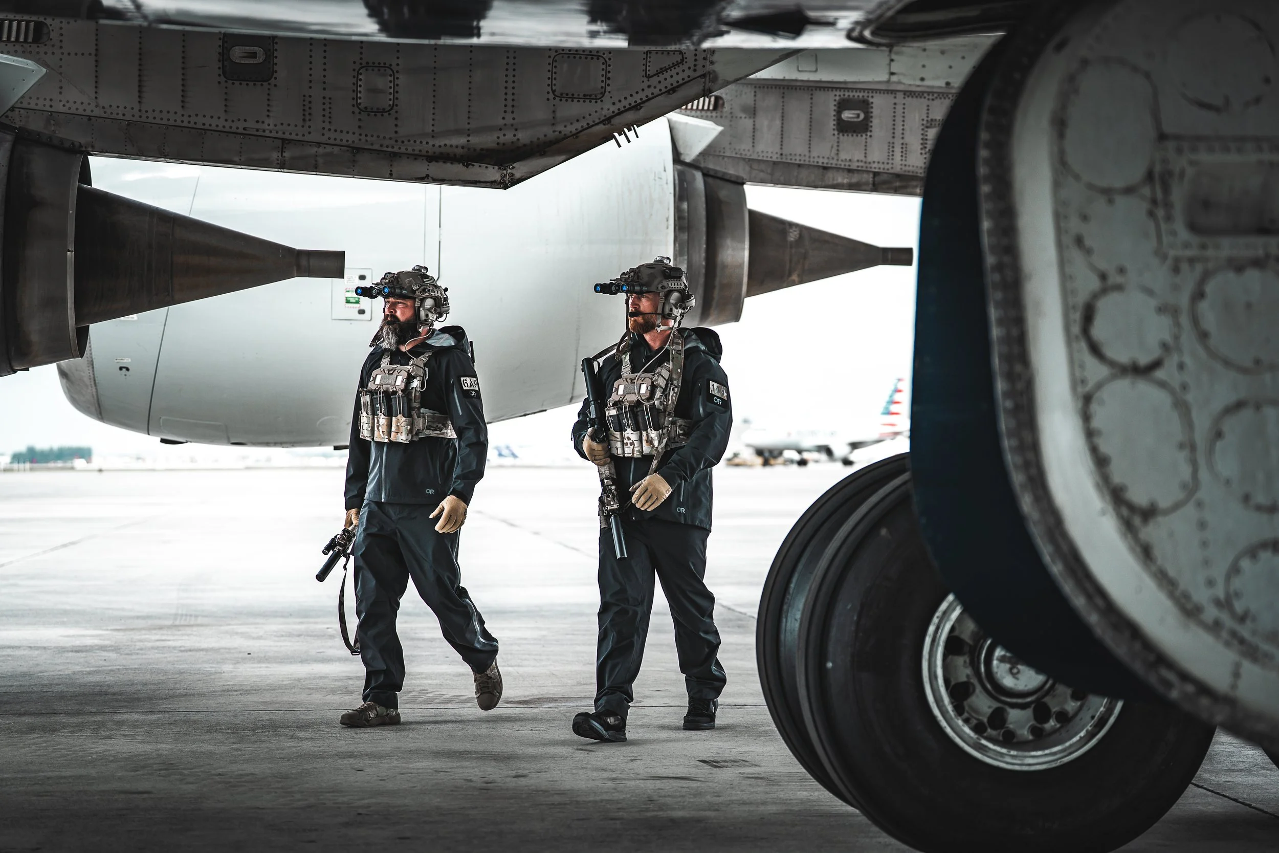 Two military personnel in tactical gear walking under a large aircraft aircraft on an airport tarmac.