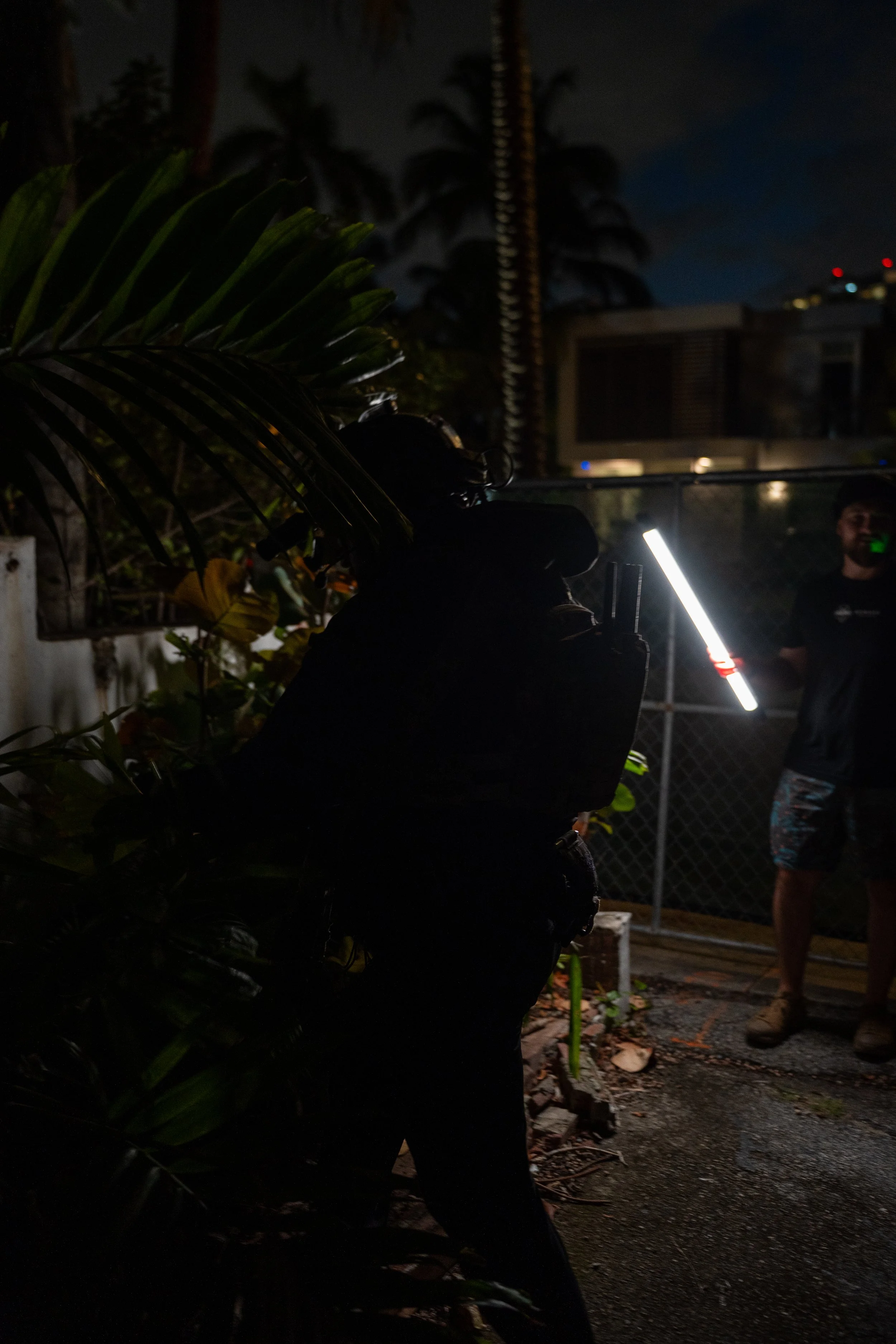 Dark silhouette of a person with a backpack and cap holding a flashlight or stick, standing in outdoor nighttime setting with dense foliage and a person in the background near a chain-link fence.