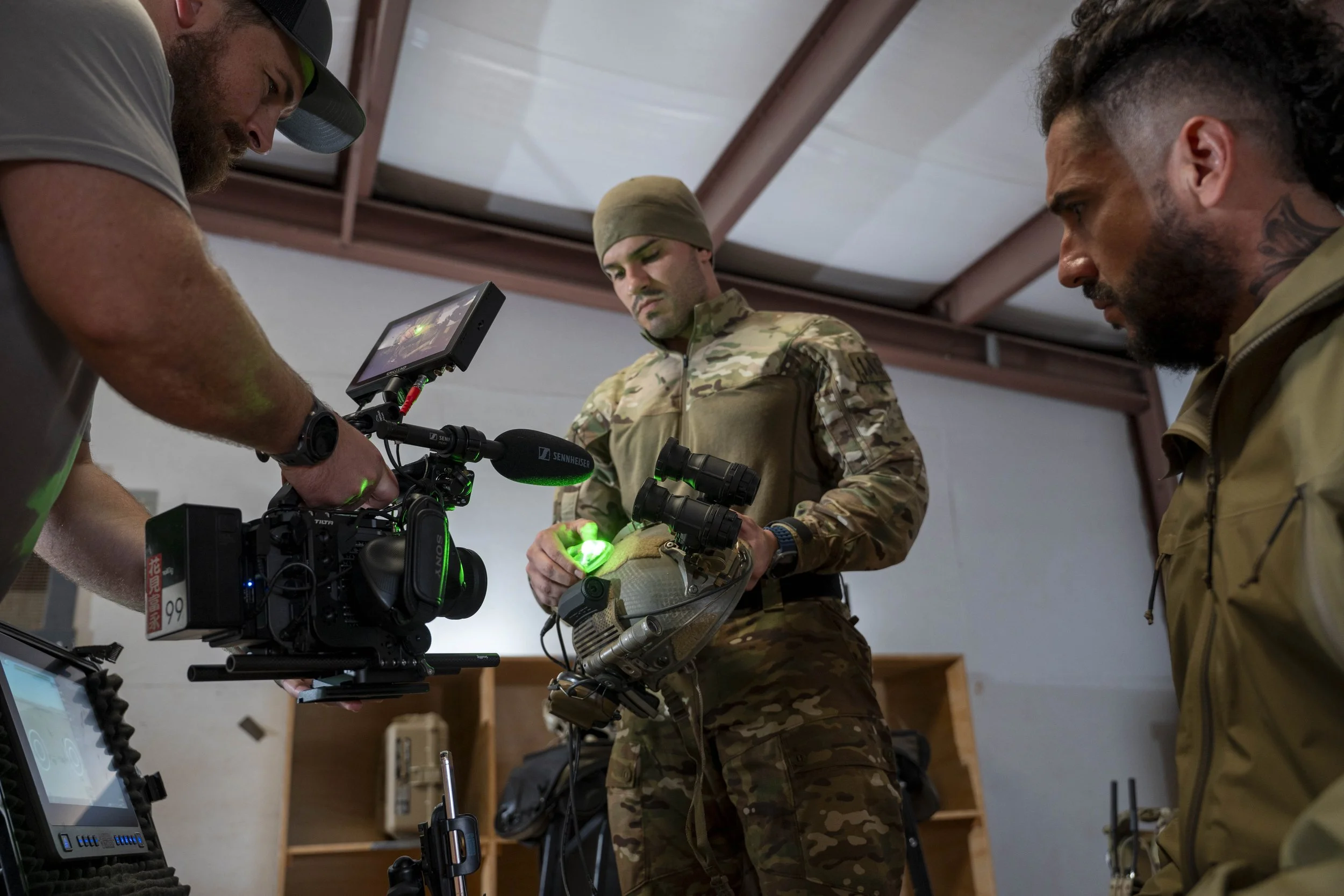 Three men, one in military camouflage, are working with filming equipment indoors. The man in the middle holds a helmet with electronic devices attached, while the other two men manage a camera and monitor.