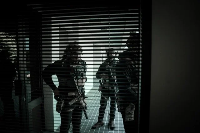 Three armed officers behind window blinds, standing in a room with a tiled floor.