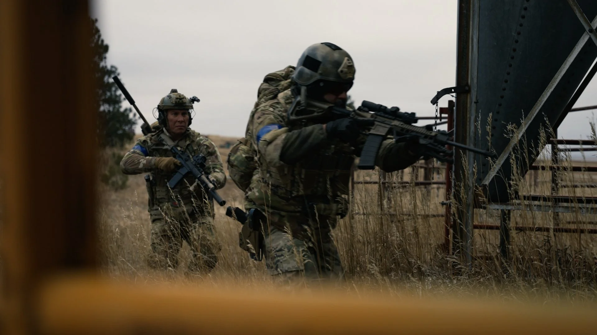 Two soldiers in camouflage uniforms and helmets, armed with rifles, moving through tall grass beside a metal structure outdoors.